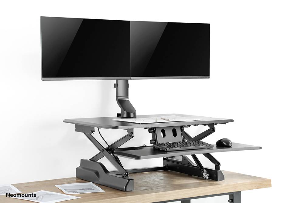Height-adjustable metal desk riser on a wooden desk with two monitors, keyboard and mouse.