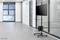An empty conference room with a large window and grey flooring. In the foreground, a mobile monitor stand with a screen is positioned.