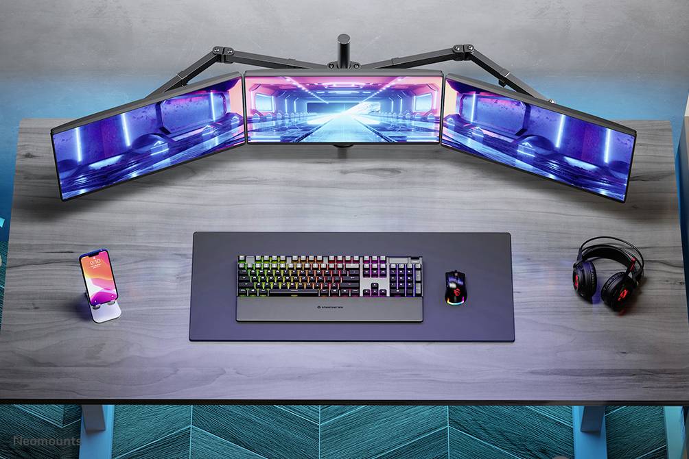 A modern desk with a keyboard, mouse, smartphone, headphones and three curved monitors displaying a futuristic blue and pink image.