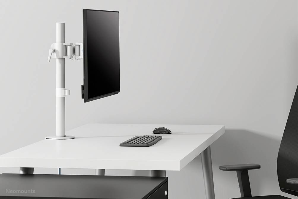 An empty office with a white desk, a monitor stand, monitor, keyboard and mouse. A black office chair stands beside it.
