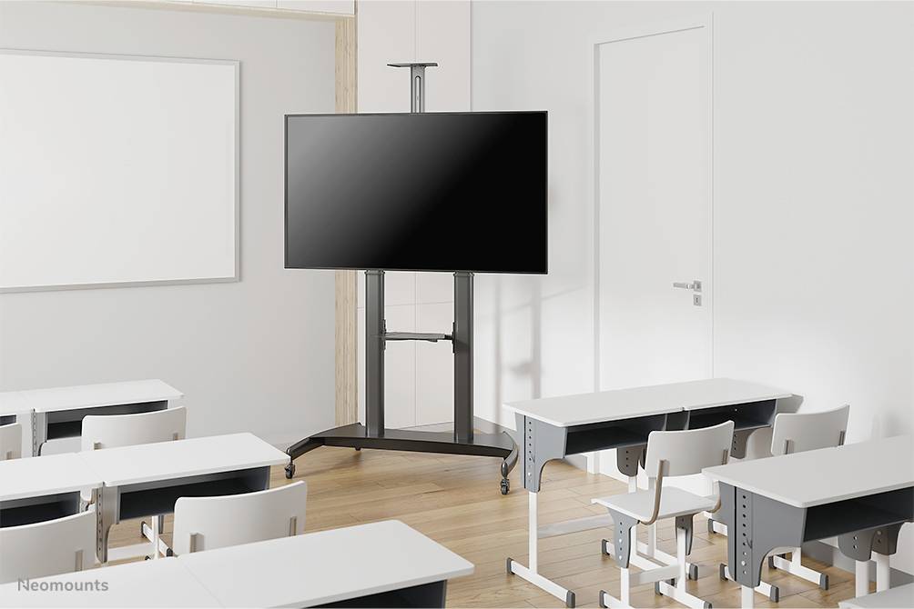 An empty classroom with white tables and chairs. A large, black screen on a mobile stand is positioned at the front next to a closed door.