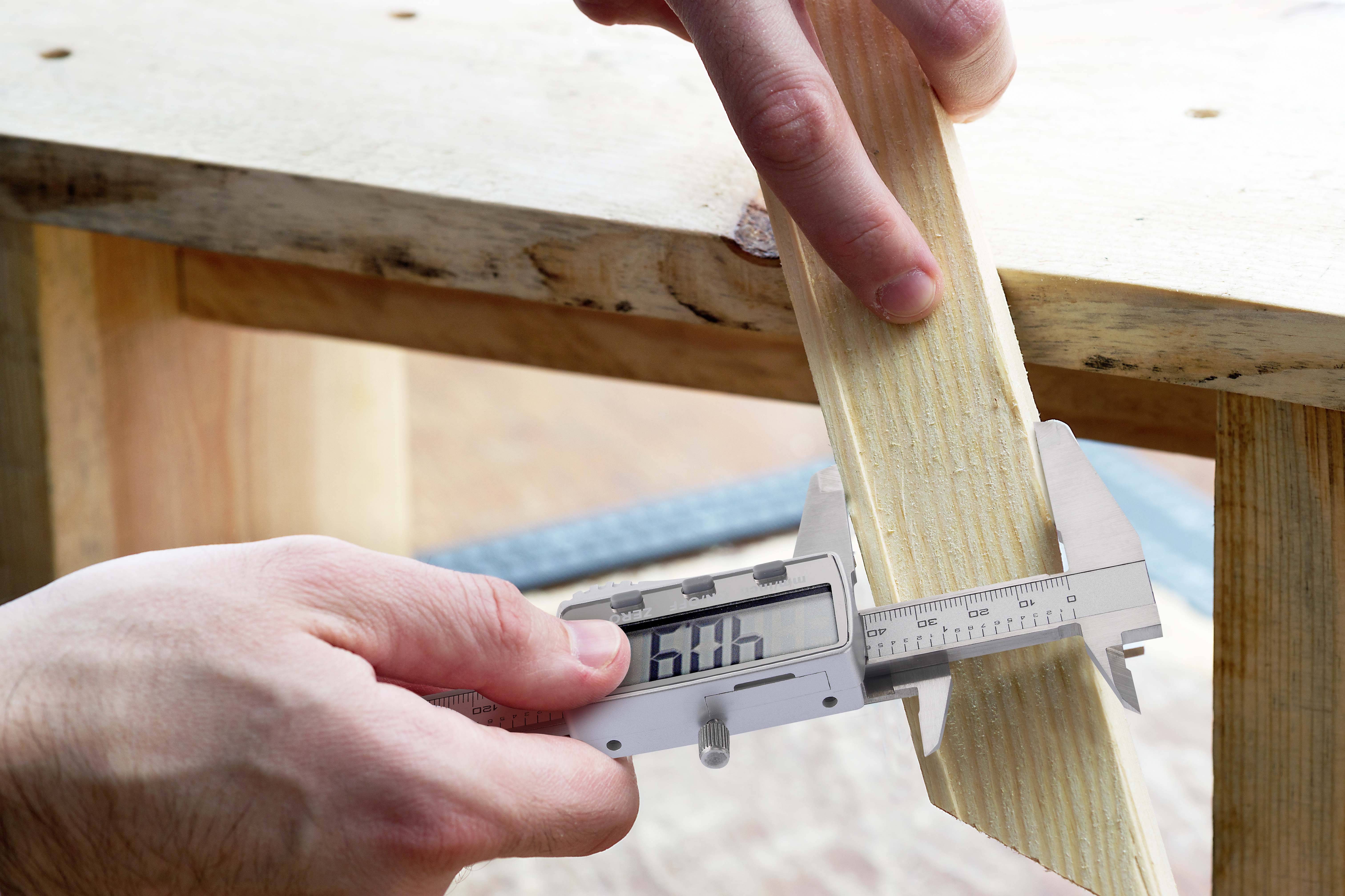 A person is measuring a wooden board with a digital calliper while holding it steady on a table.