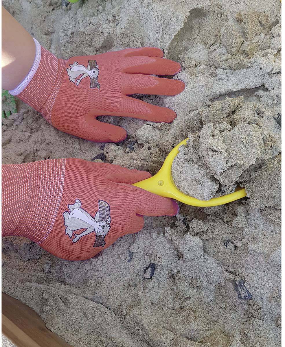 A child digs in the sand with a yellow spade, wearing red gloves with a rabbit design.