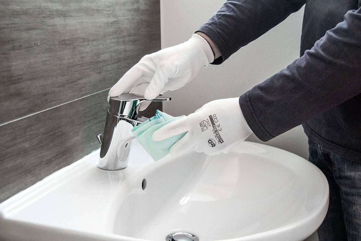 A person is cleaning a sink tap with a cloth, wearing white gloves.