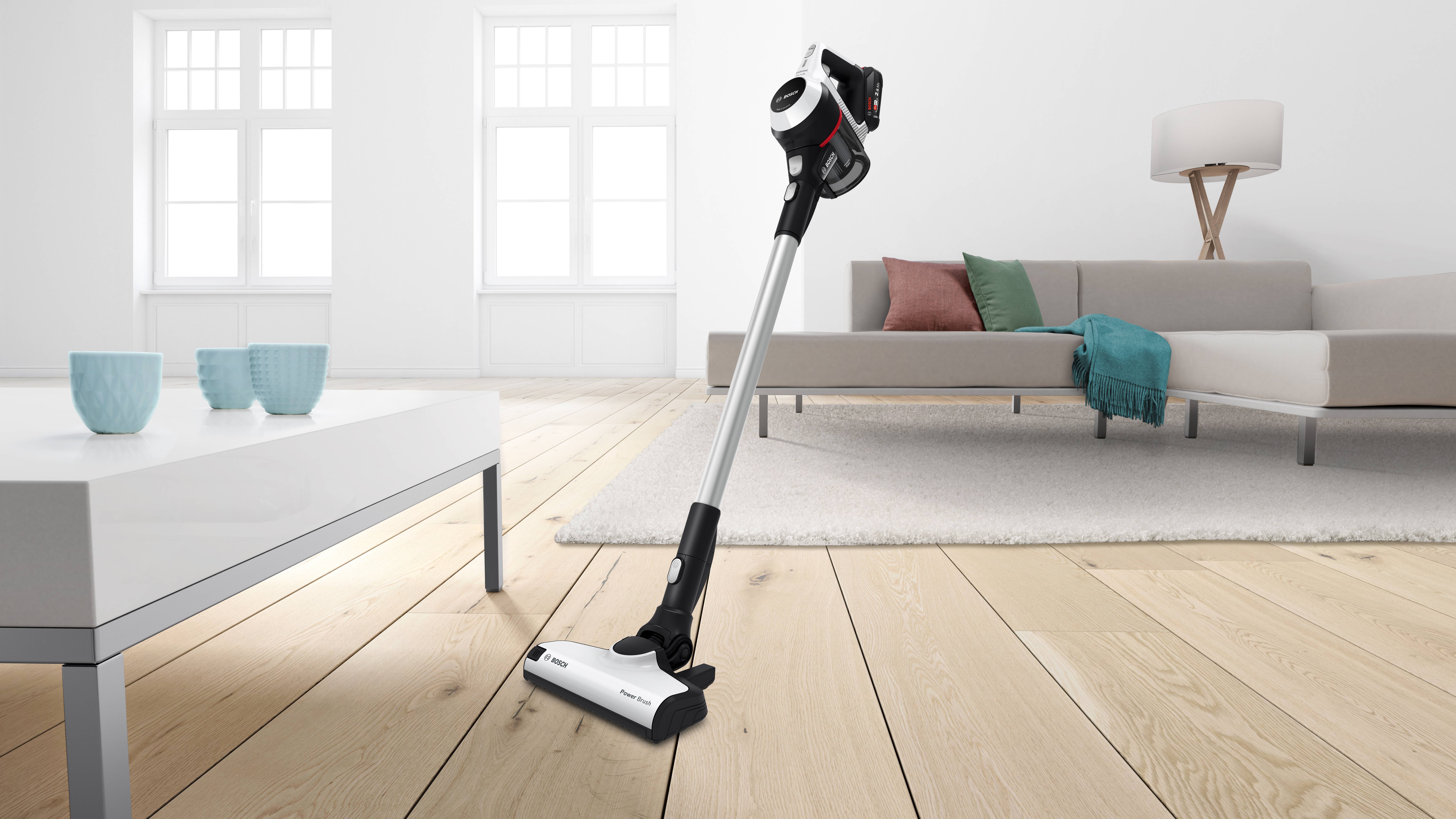 Cordless vacuum cleaner stands on a wooden floor in a modern living room with a sofa, table, and lamp. Bright and minimalist interior.