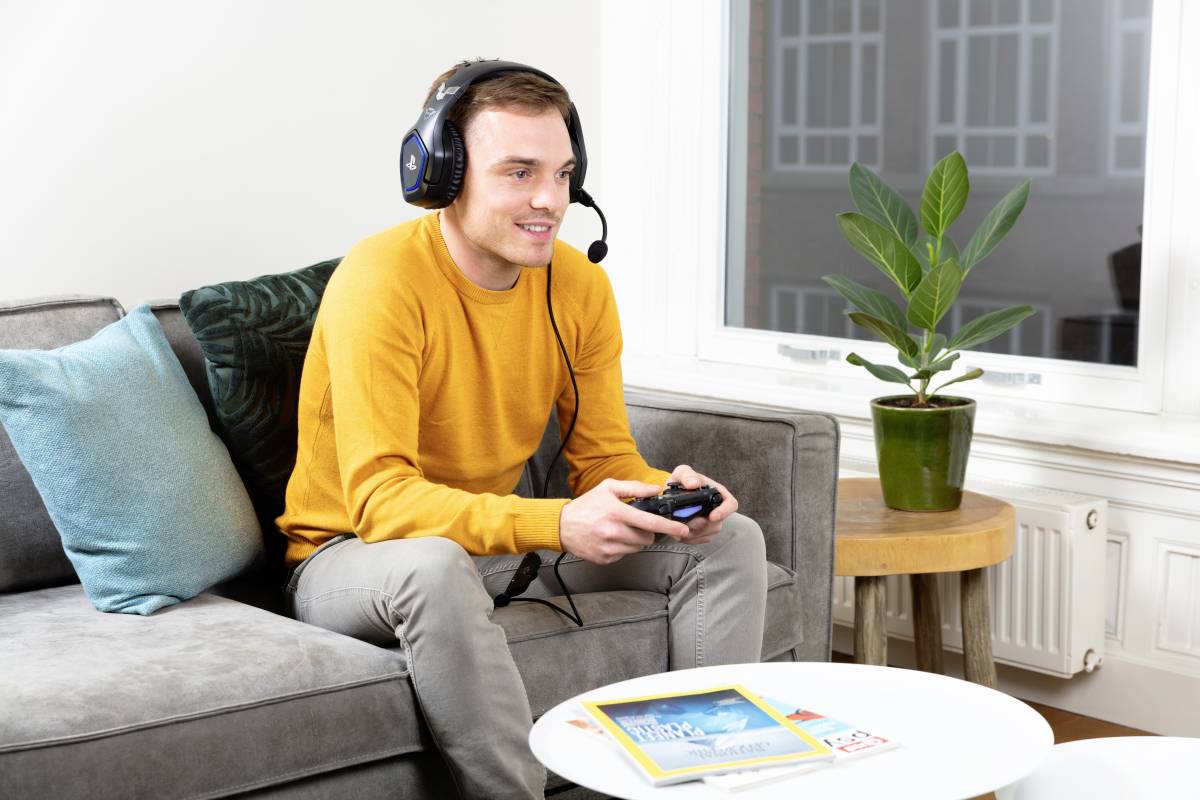 A man is sitting on a sofa and playing with a controller while wearing a headset. A magazine is lying on the table beside him.
