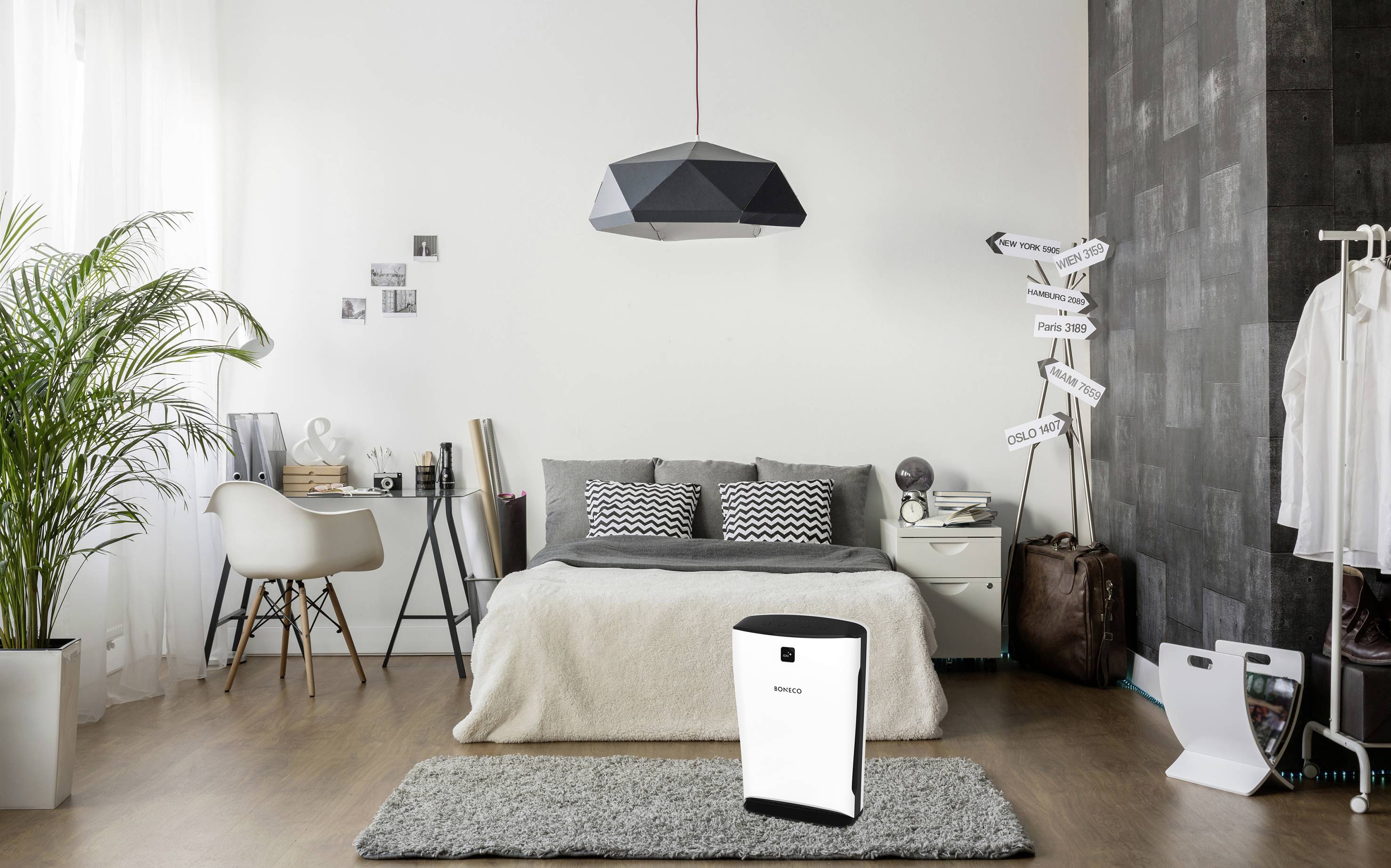 Bedroom with a bed, desk, and plants. A white air purifier sits on the carpet. Bright walls, modern pendant light.