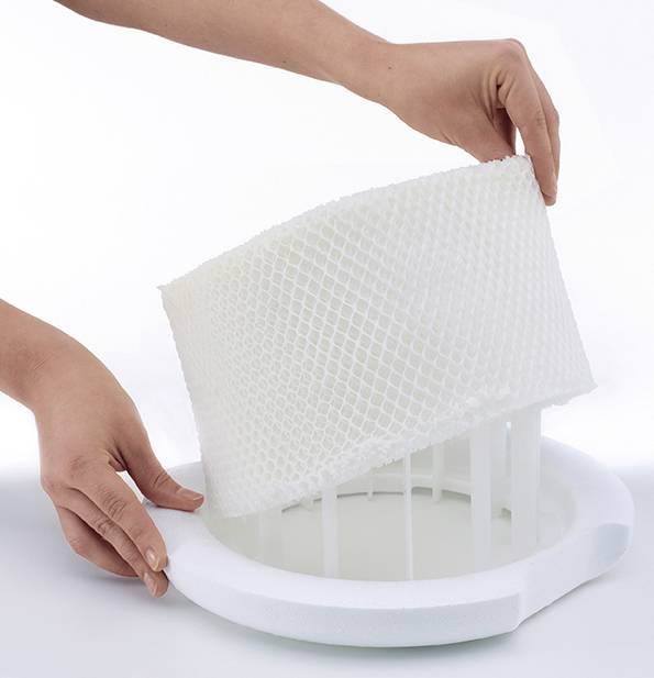 Two hands are removing a white filter from an air purifier. The filter has a honeycomb-like structure and is cylindrical in shape.