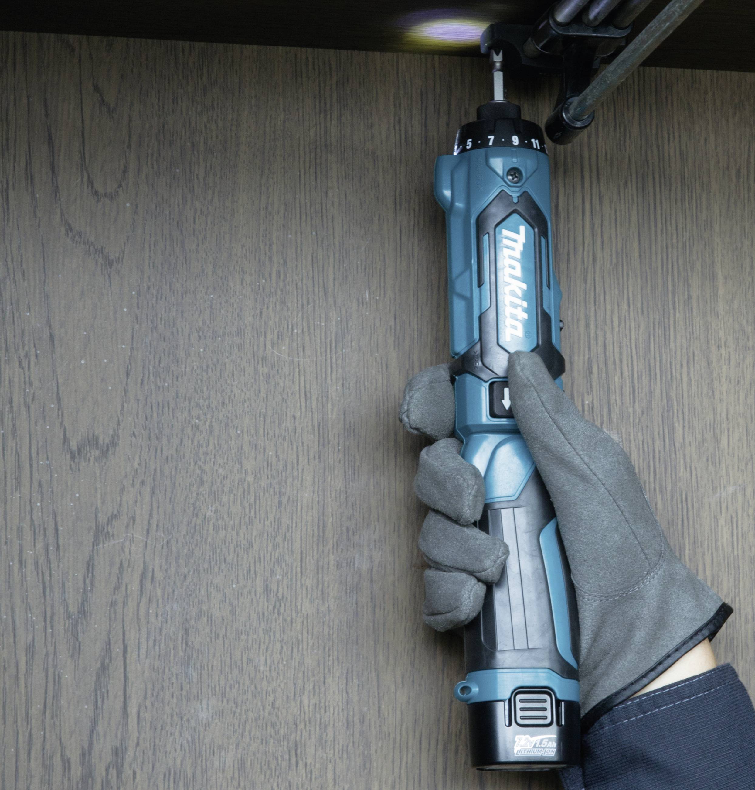 A person tightens a screw with an electric screwdriver. The hand is wearing a grey glove.