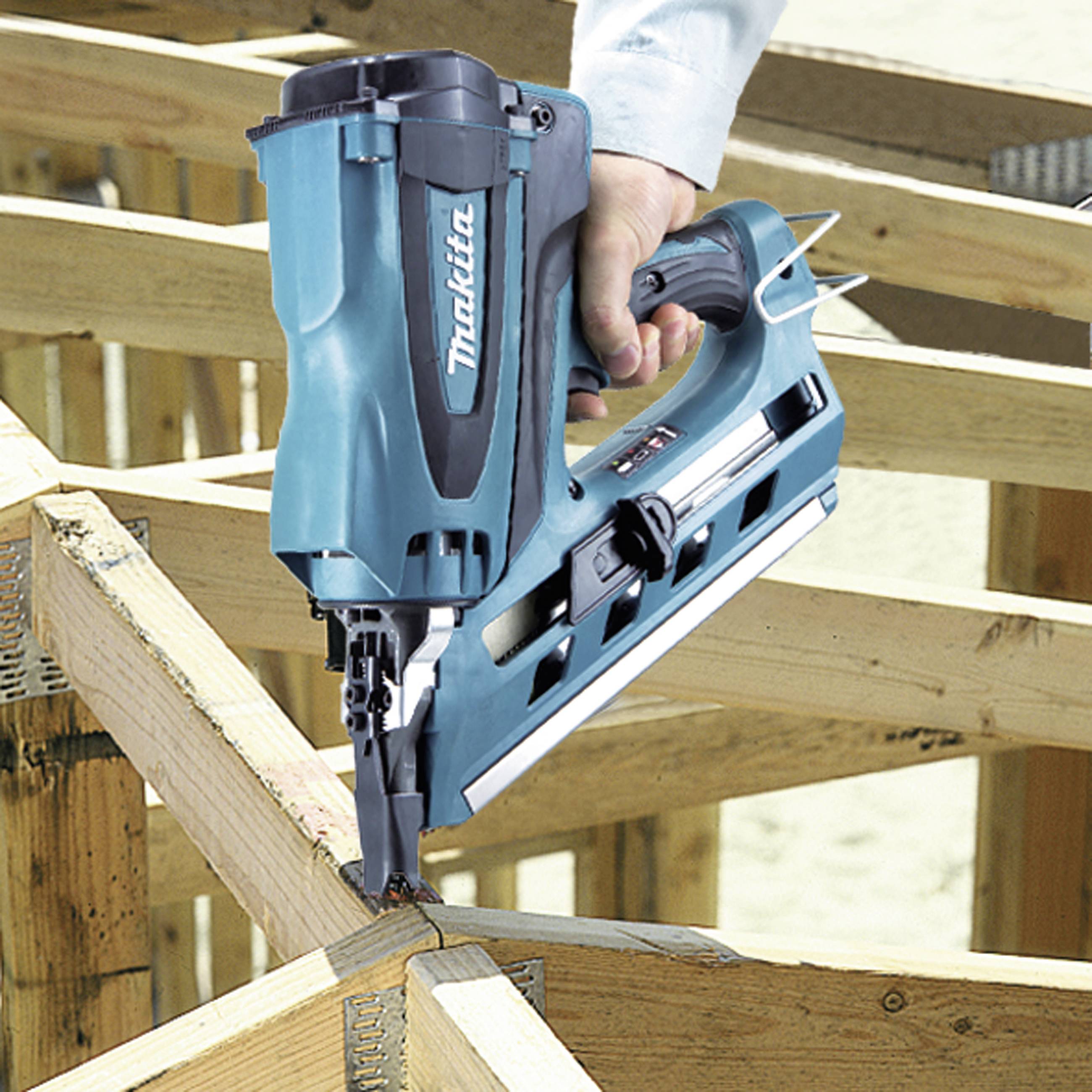 A person is working with a blue cordless nail gun on a wooden frame construction. The nail gun is being pressed against a piece of wood.