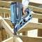 A person is working with a blue cordless nail gun on a wooden frame construction. The nail gun is being pressed against a piece of wood.