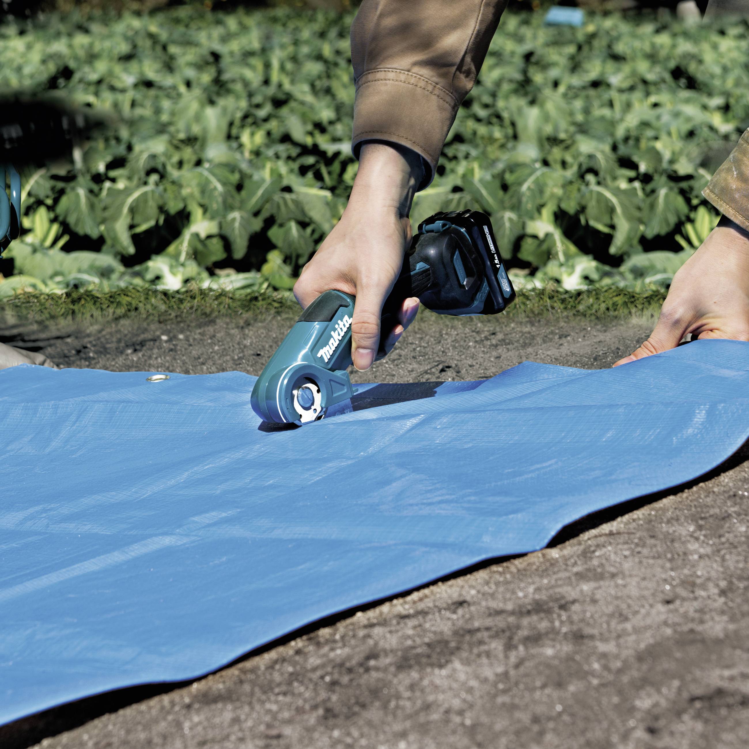 A person is cutting a blue tarpaulin in the garden using a cordless tool. Green plants can be seen in the background.