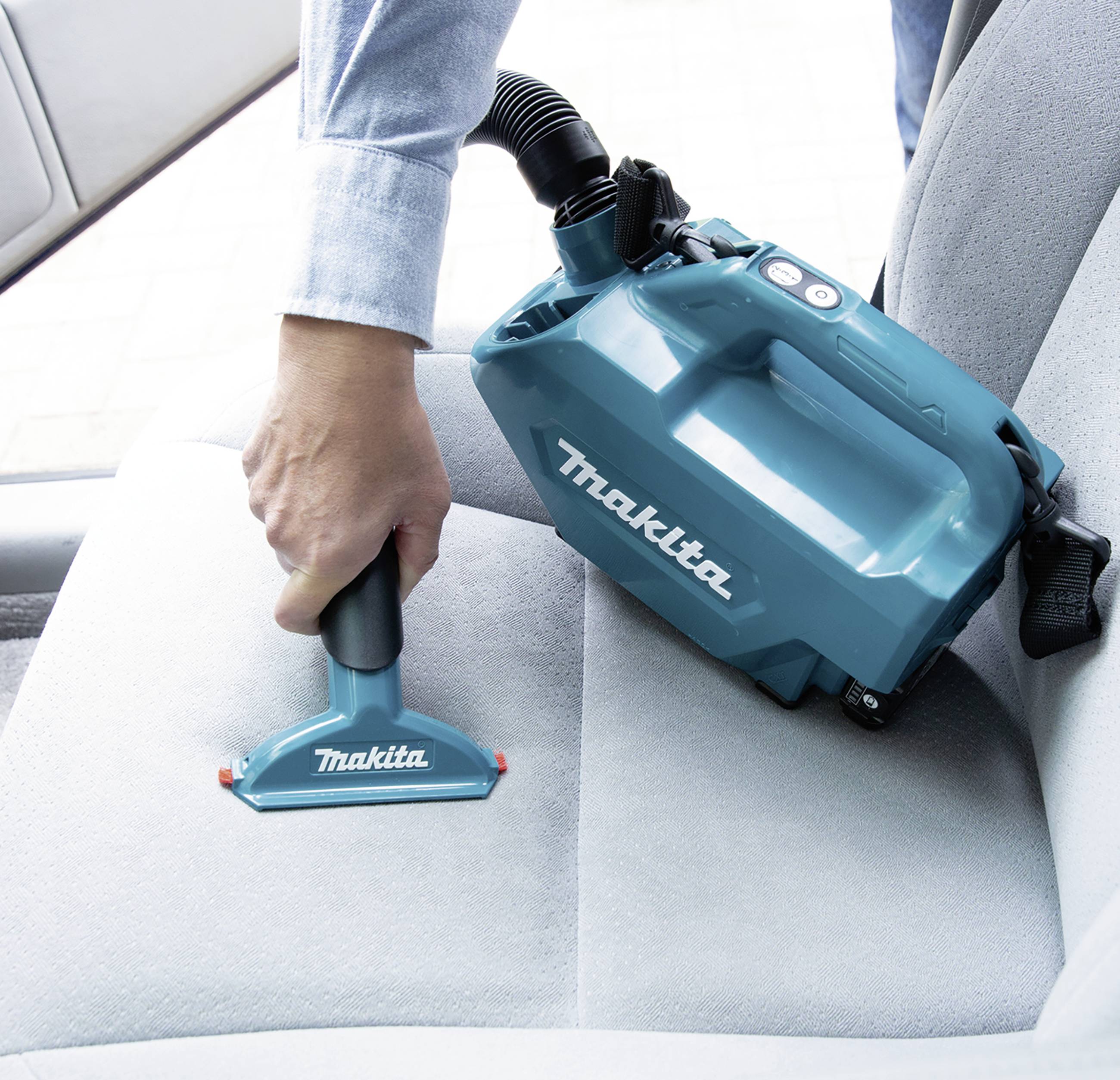 A person is vacuuming a car seat with a blue, portable Makita handheld vacuum. The vacuum is being held with one hand.