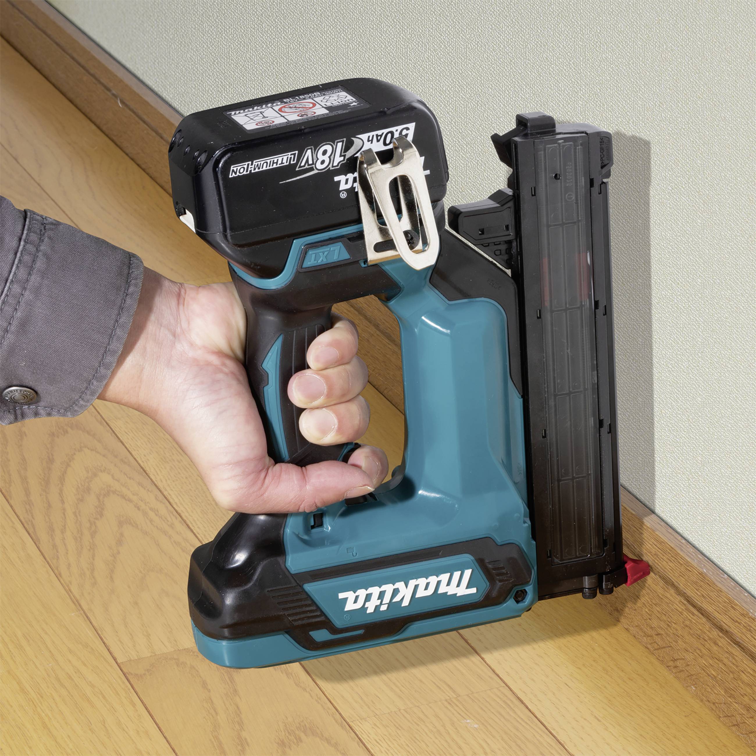 A person is holding a blue cordless nail gun and securing a black strip to a wall. The tool has a brand logo printed on it.