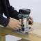 A person is operating a hand router on a wooden table, likely as part of a woodworking task.