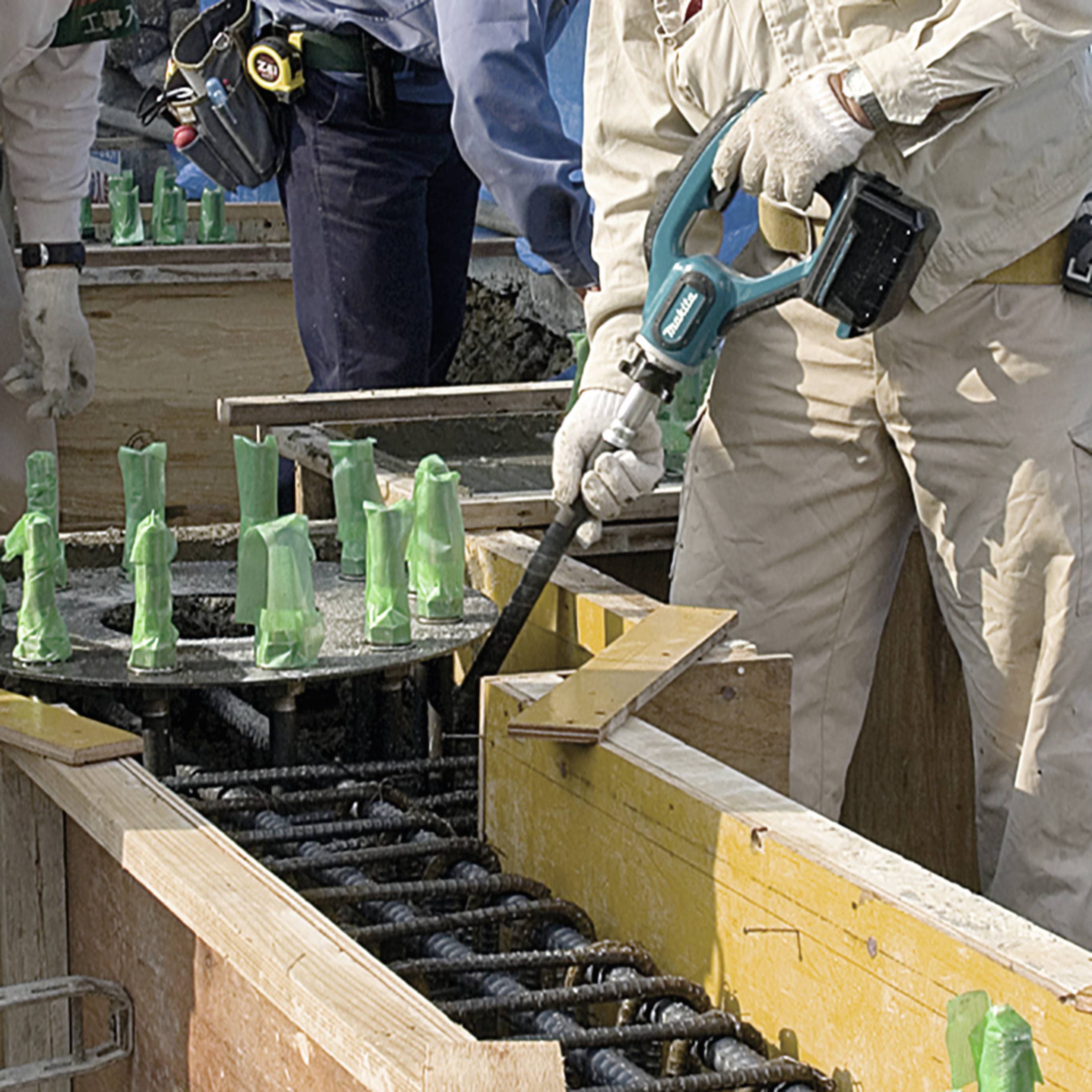 Construction workers are pouring concrete into a wooden formwork secured with green reinforcements. One person is holding a vibration compactor.
