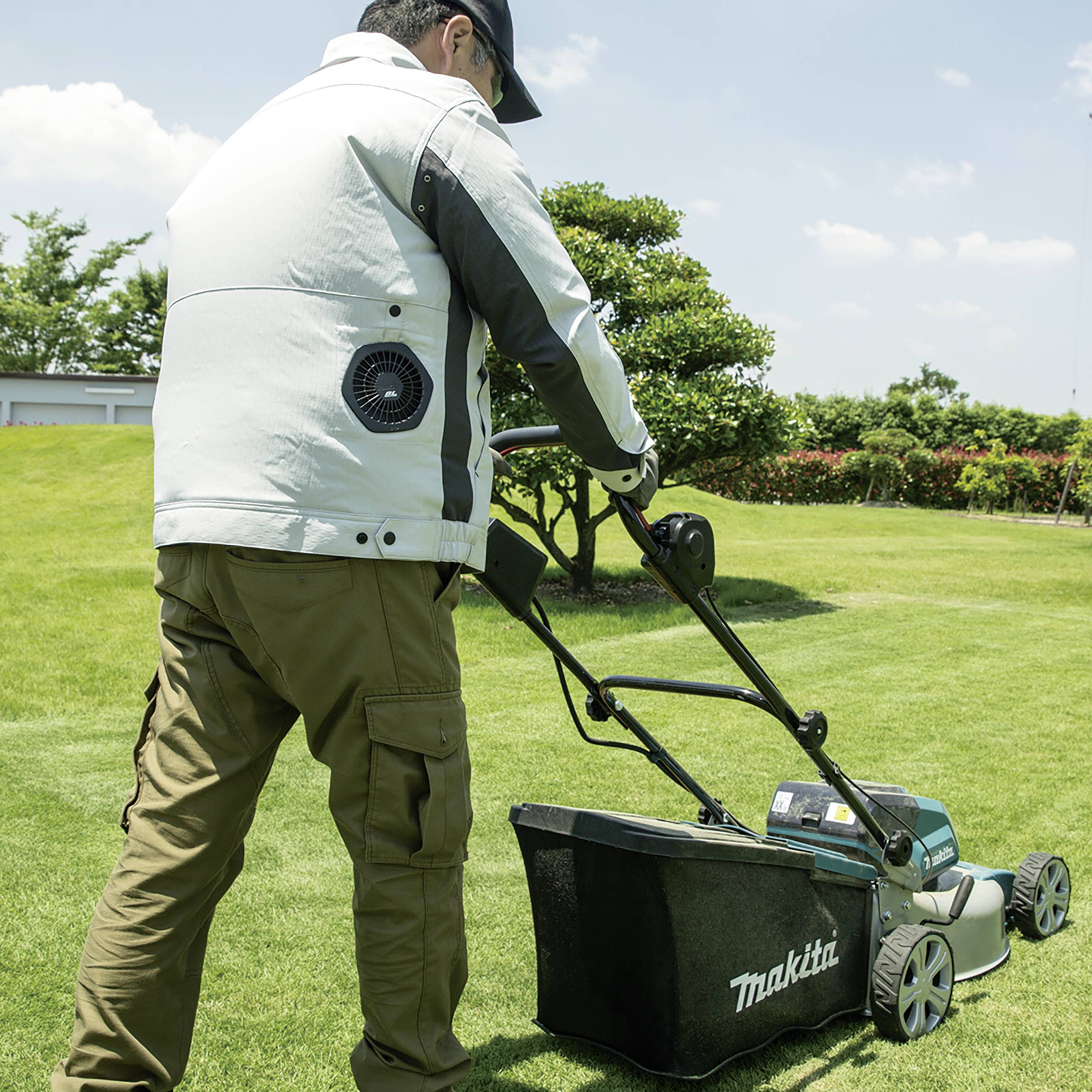 A person is pushing a lawnmower across a green lawn. Trees and bushes in the background, on a sunny day.