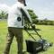 A person is mowing the lawn with an electric lawnmower in a garden. The sky is clear and there are trees in the background.