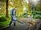 A man is using a leaf blower in an autumnal garden. Leaves are being blown away from a pathway. Trees and shrubs are visible in the background.