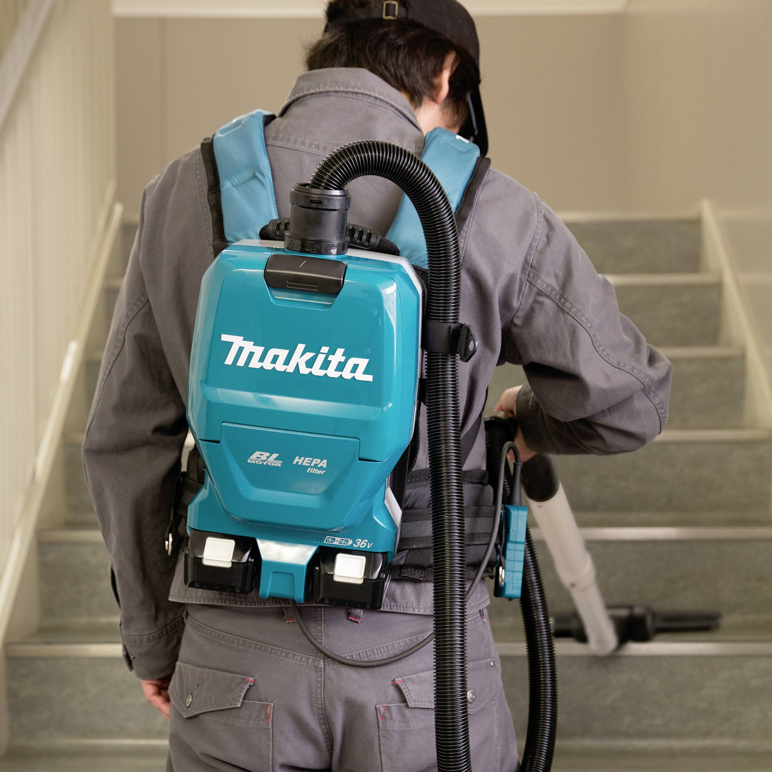 A person is cleaning a staircase with a backpack vacuum cleaner. The vacuum cleaner is light blue and bears the 'Makita' logo.