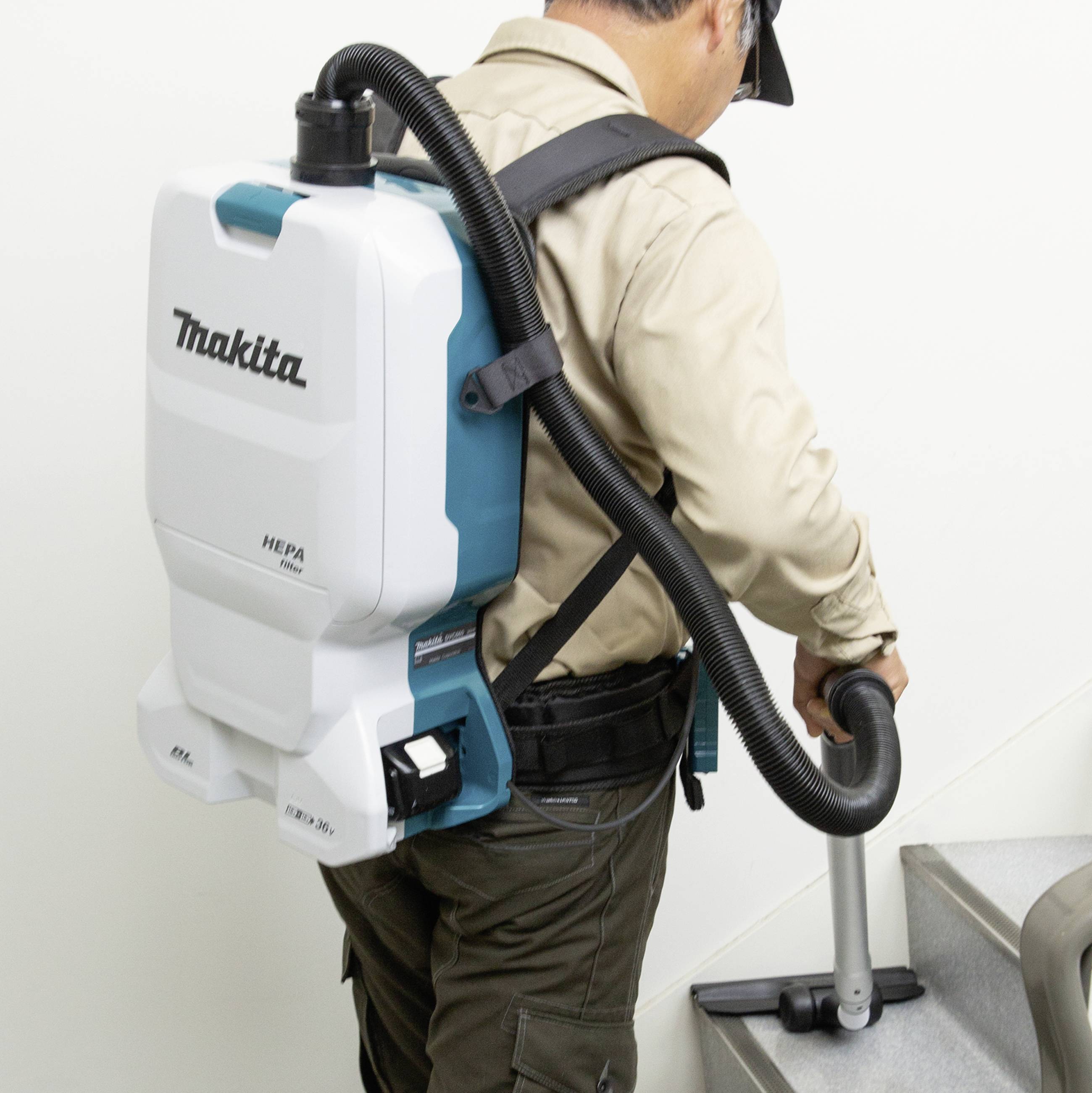 A person is using a portable vacuum cleaner on a staircase. The vacuum cleaner is attached to their back and is manufactured by Makita.