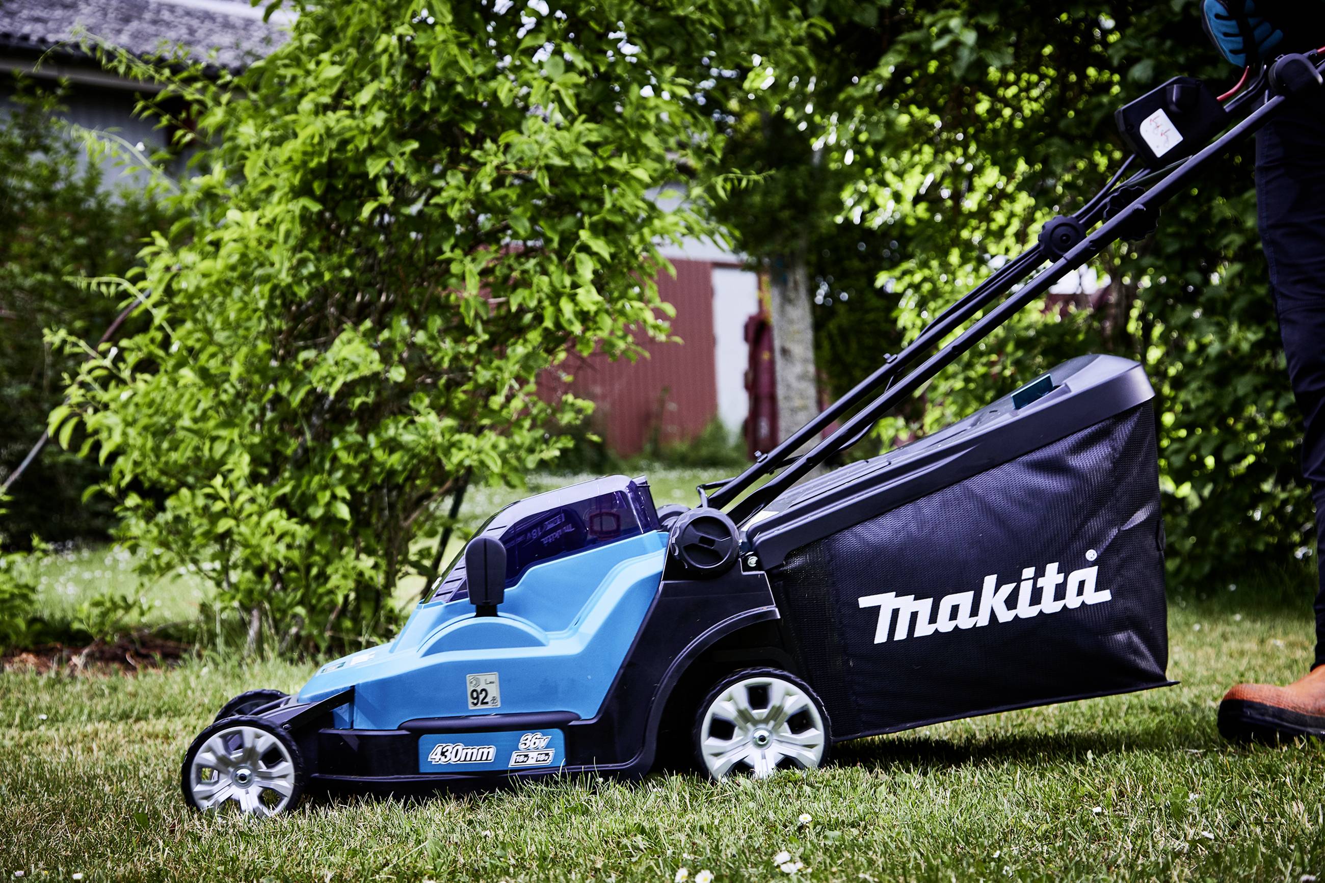A blue lawnmower with a 'Makita' logo stands on a green lawn. A person is pushing the mower. Bushes are visible in the background.