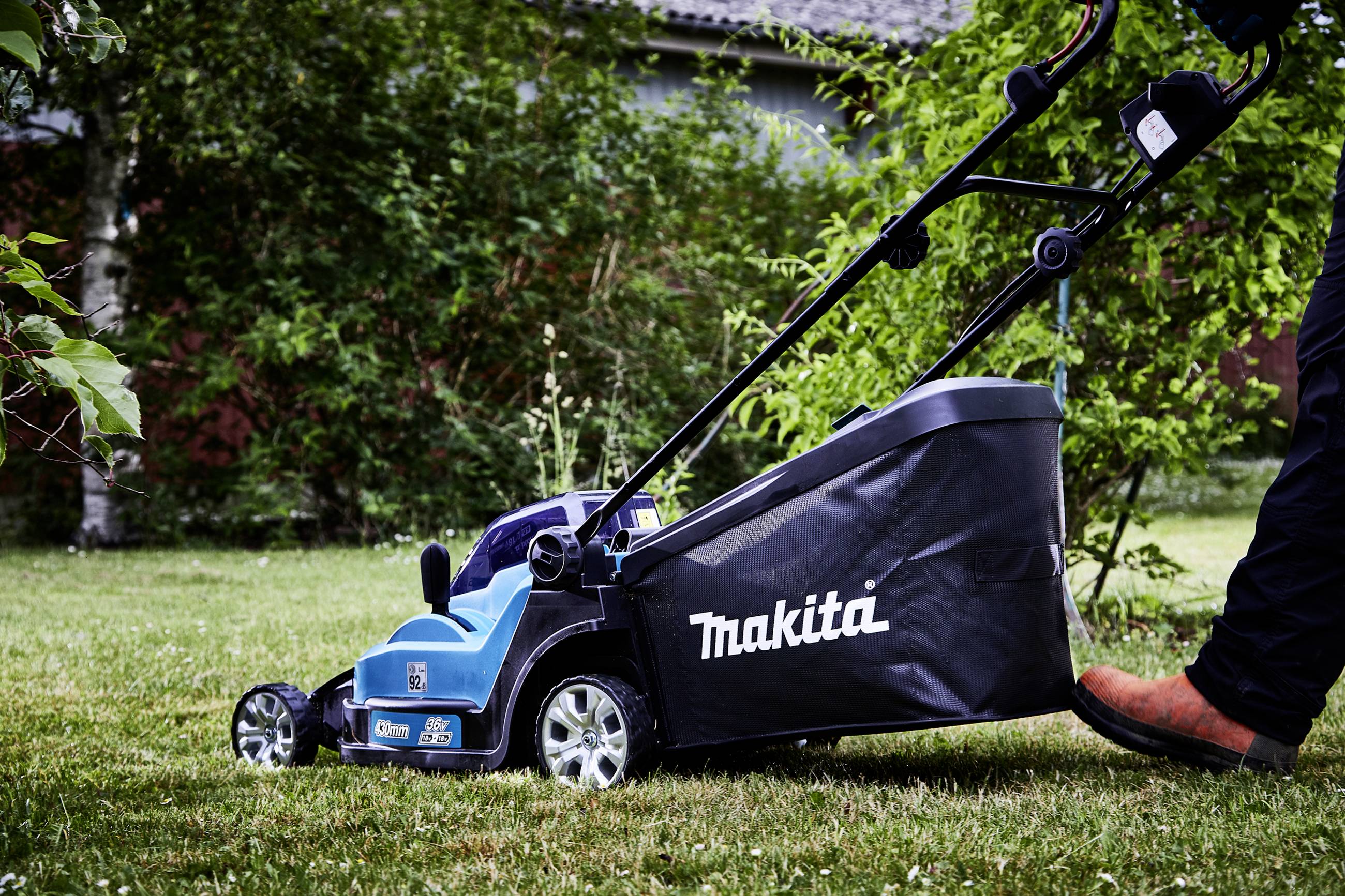 A lawnmower is being pushed by a person across a green lawn. The collection bag of the lawnmower is marked with 'Makita'. A garden can be seen in the background.
