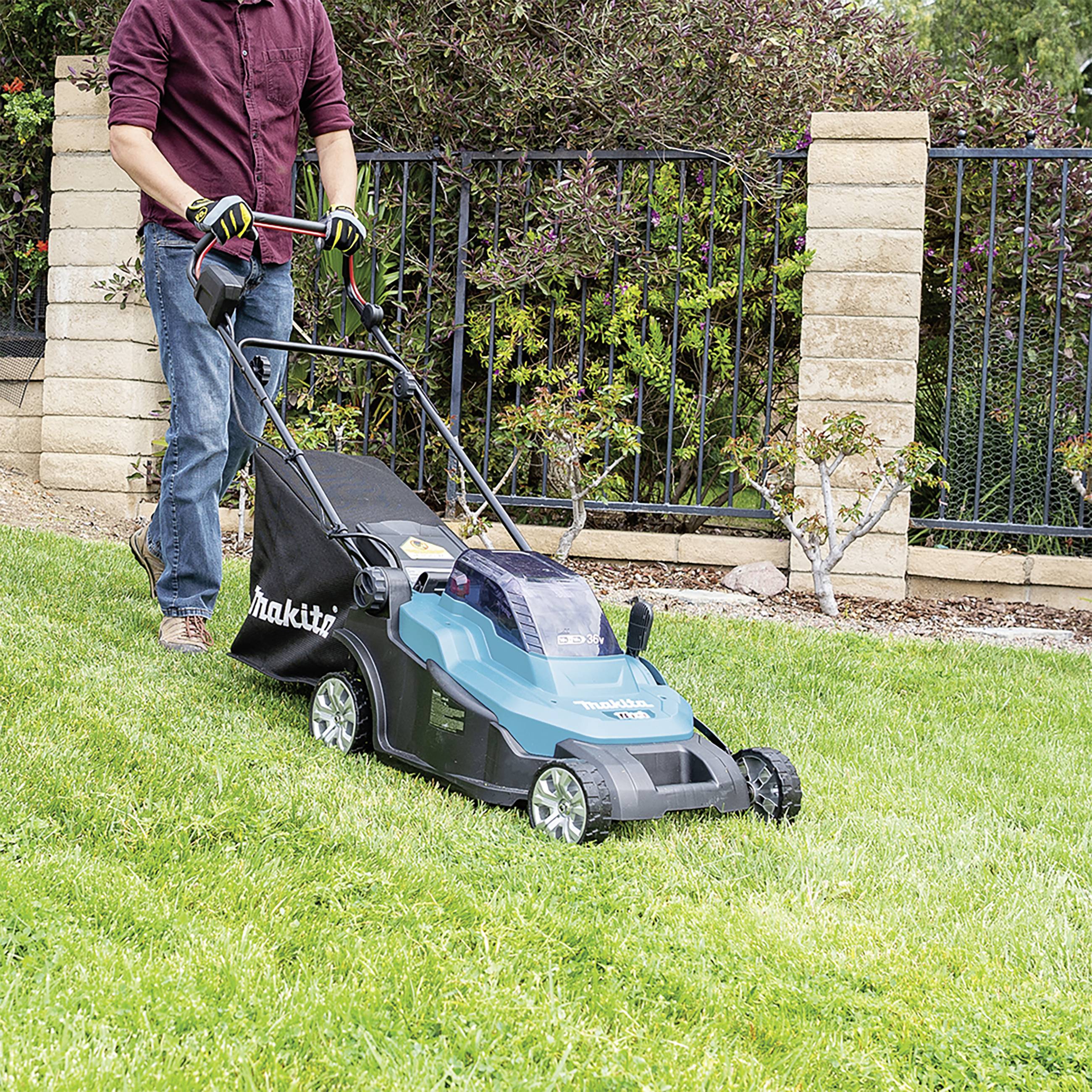 A person is mowing a lawn with an electric lawnmower in a garden with grassy areas and ornamental plants in the background.