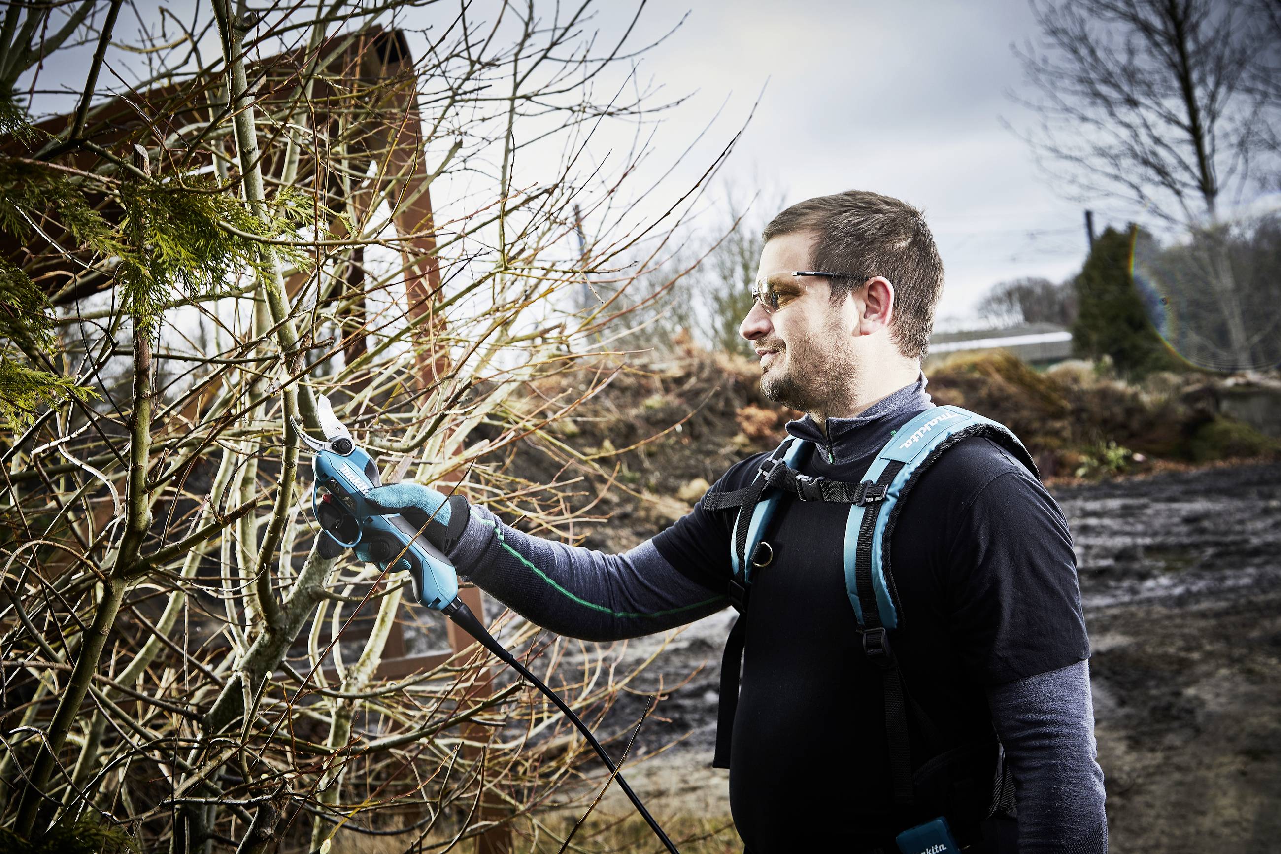 A man wearing safety glasses is cutting branches with an electric pruning saw on a tree. A landscape is visible in the background.