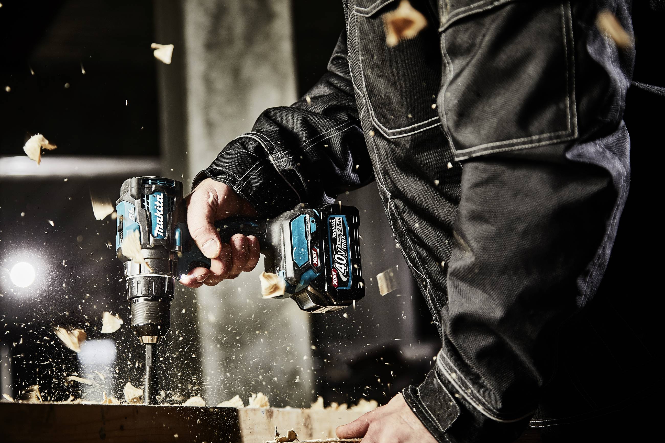 A person in black work attire is drilling into wood with an electric drill, wood shavings flying into the air.