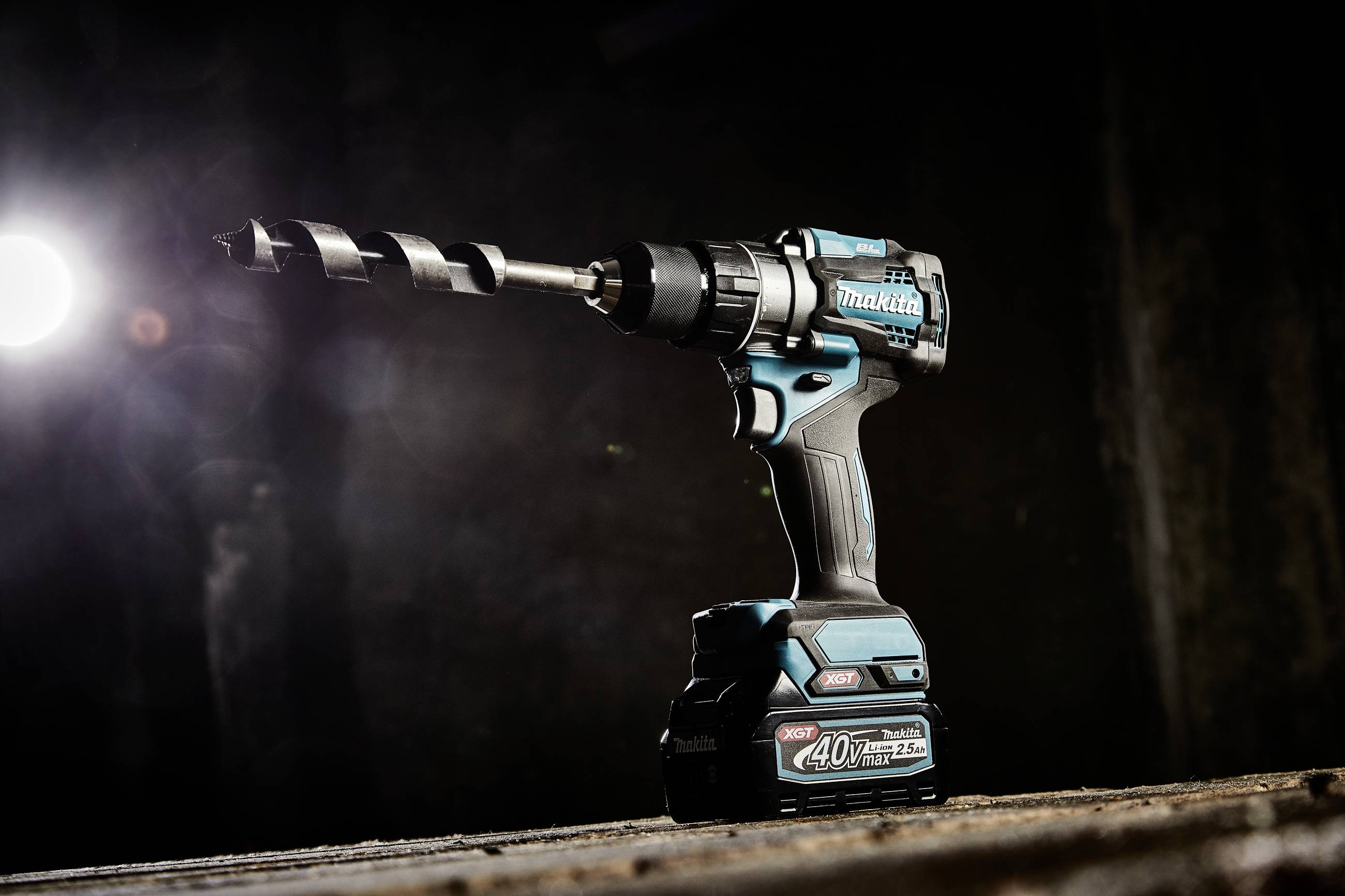 'Cordless drill with large drill bit, illuminated from the left, on a workbench in a dimly lit environment.'