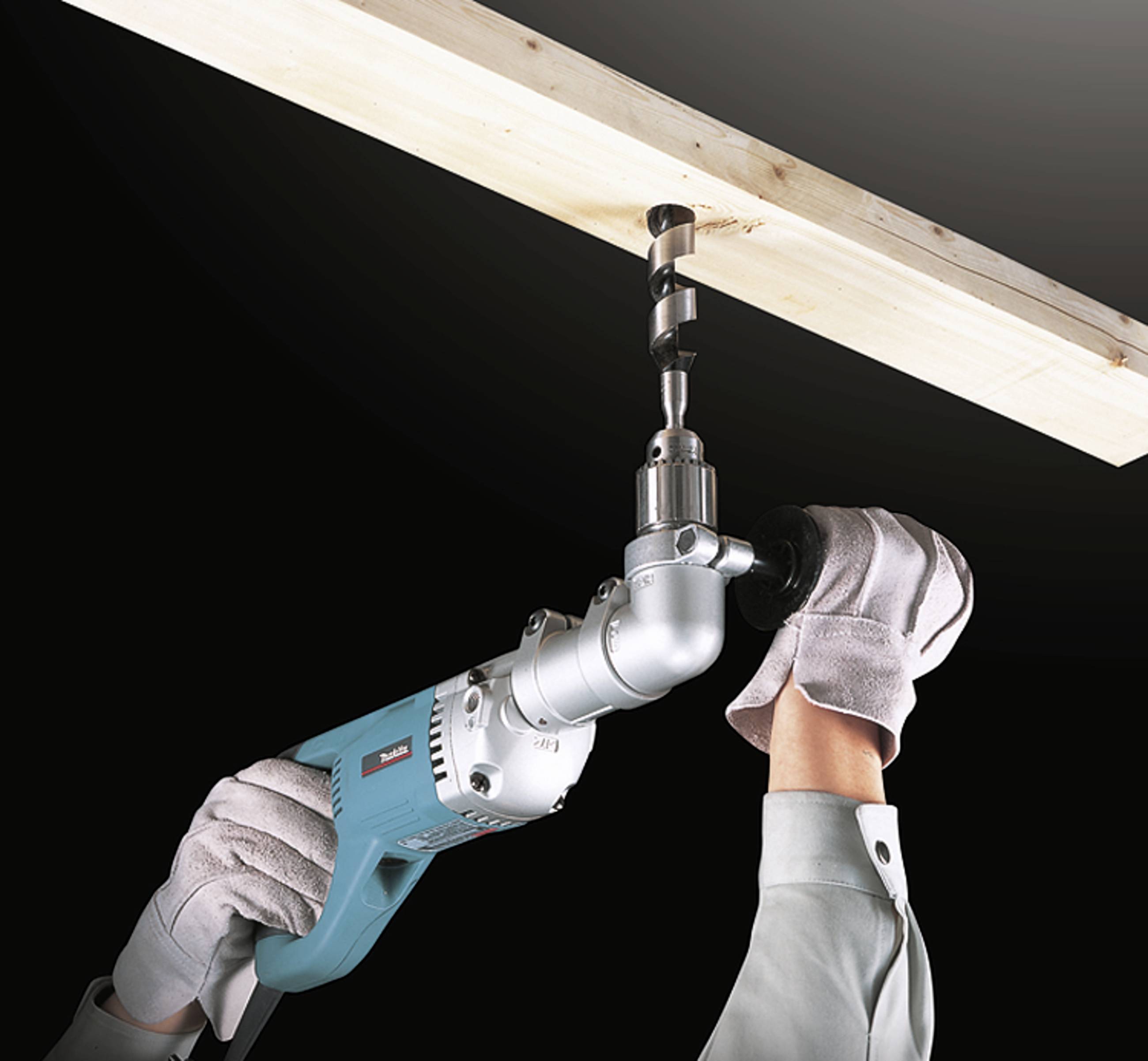 A person is drilling a hole in a wooden board using a blue electric tool. The person is wearing gloves for safety.