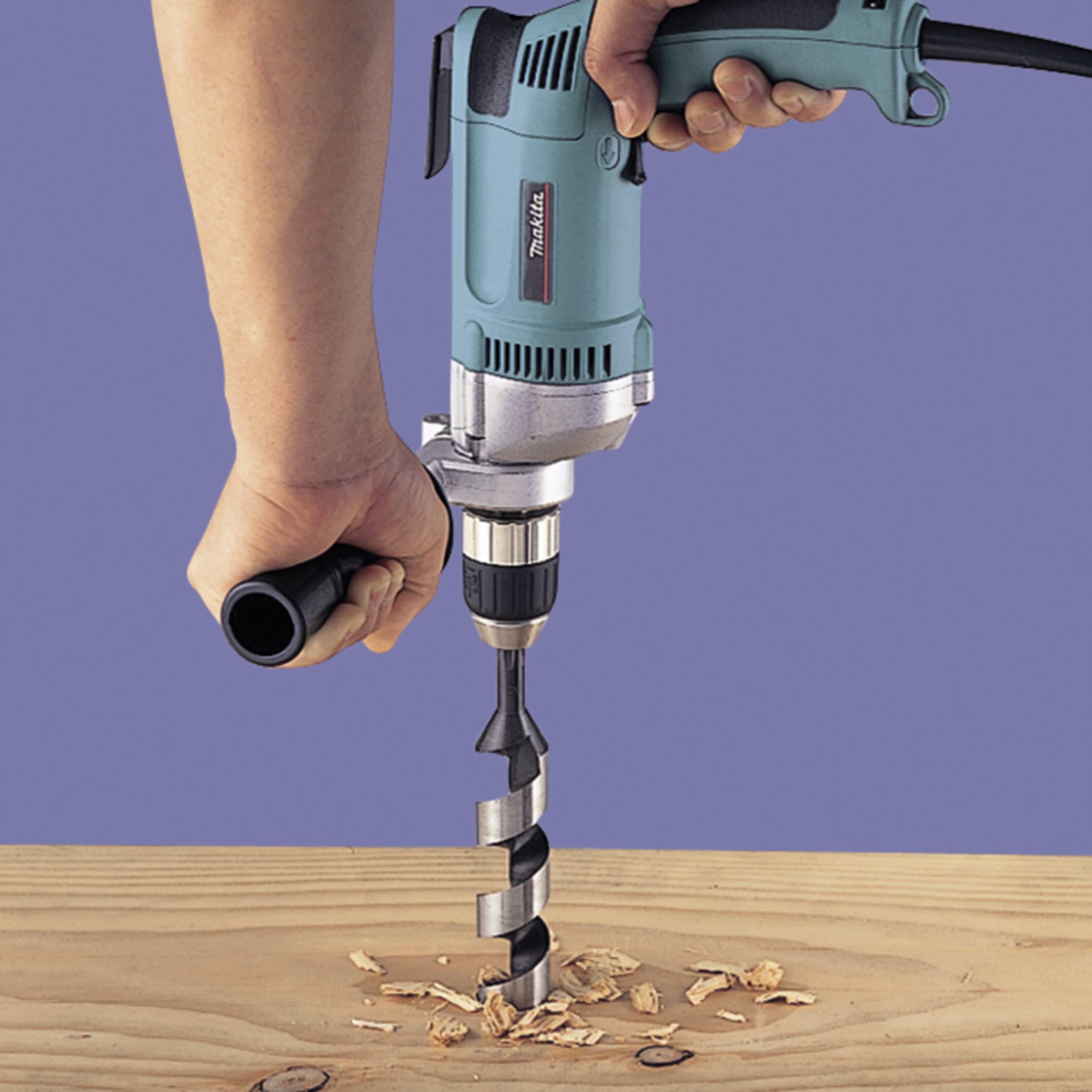A hand is holding a drill, which is boring into a wooden board. Wood shavings are scattered around the drill hole, in a workshop environment.