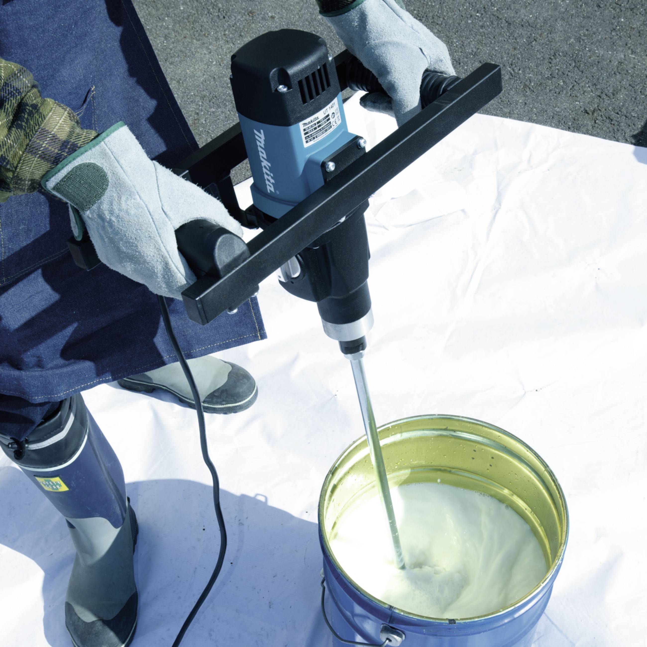 A person is mixing white paint in a bucket using an electric mixing device. Protective clothing and gloves are being worn.
