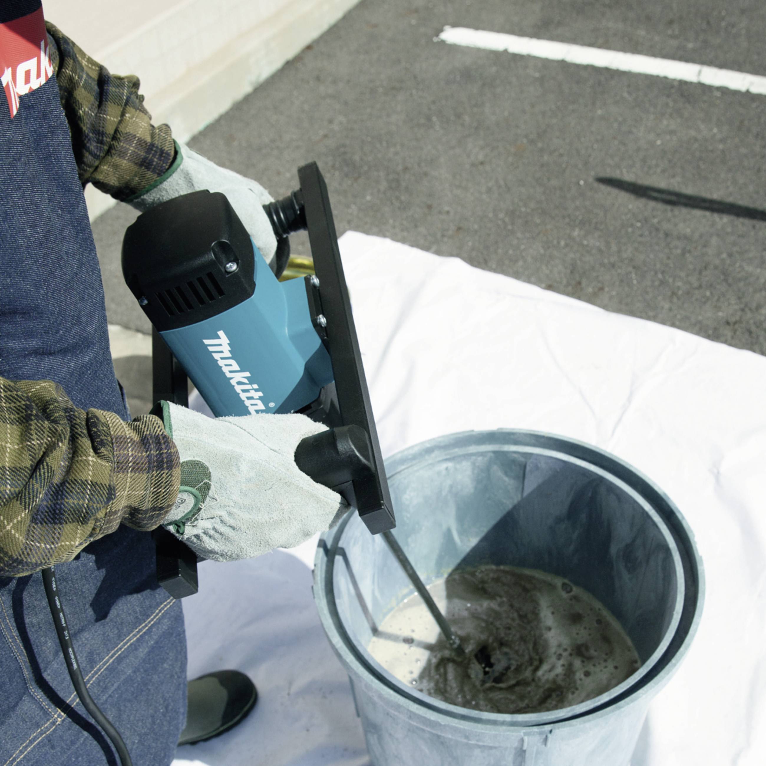 'Person in work attire mixing liquid in a bucket with an electric drill outdoors.'