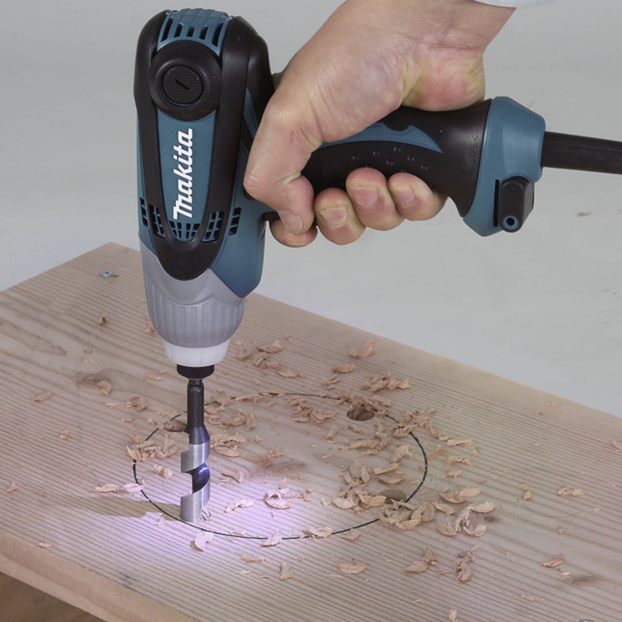 A hand holds a cordless drill and is drilling a hole into a wooden board. Sawdust is scattered around the hole.