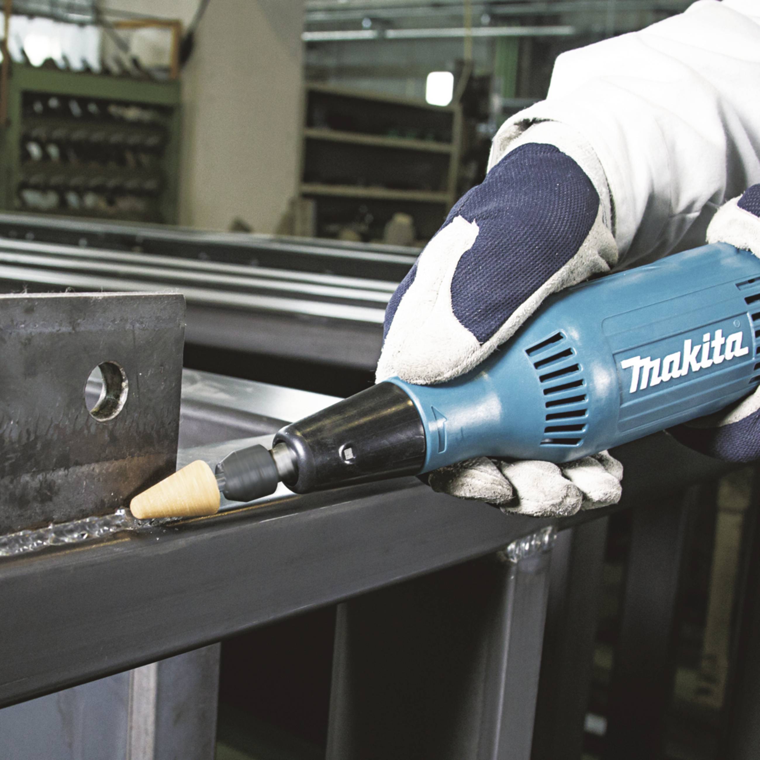 A person wearing protective gloves is holding a 'Makita' angle grinder and working on a metal surface in a workshop environment.