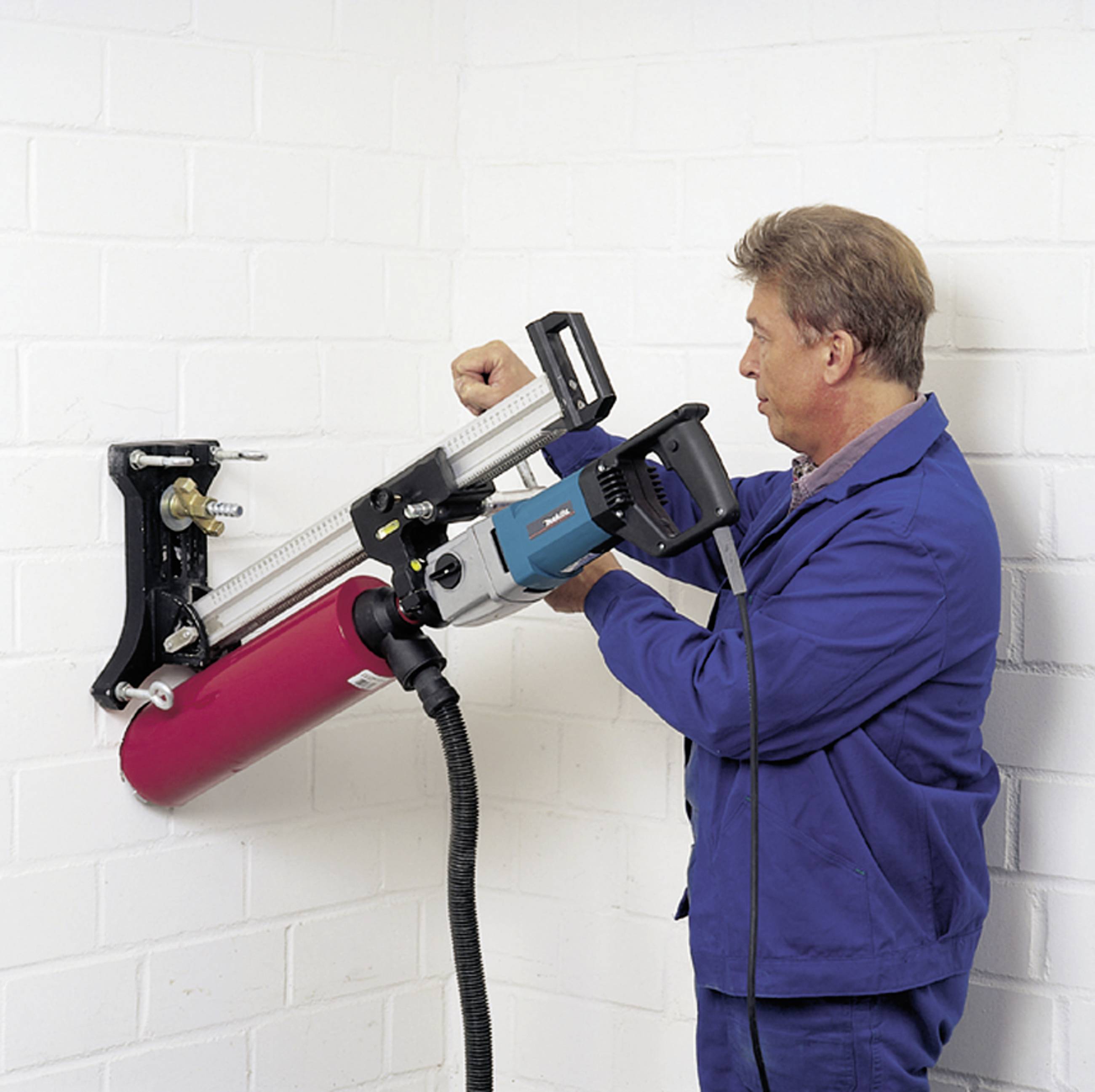 A tradesman is drilling into a white brick wall with a large machine. He is wearing blue workwear and is focused on the task.