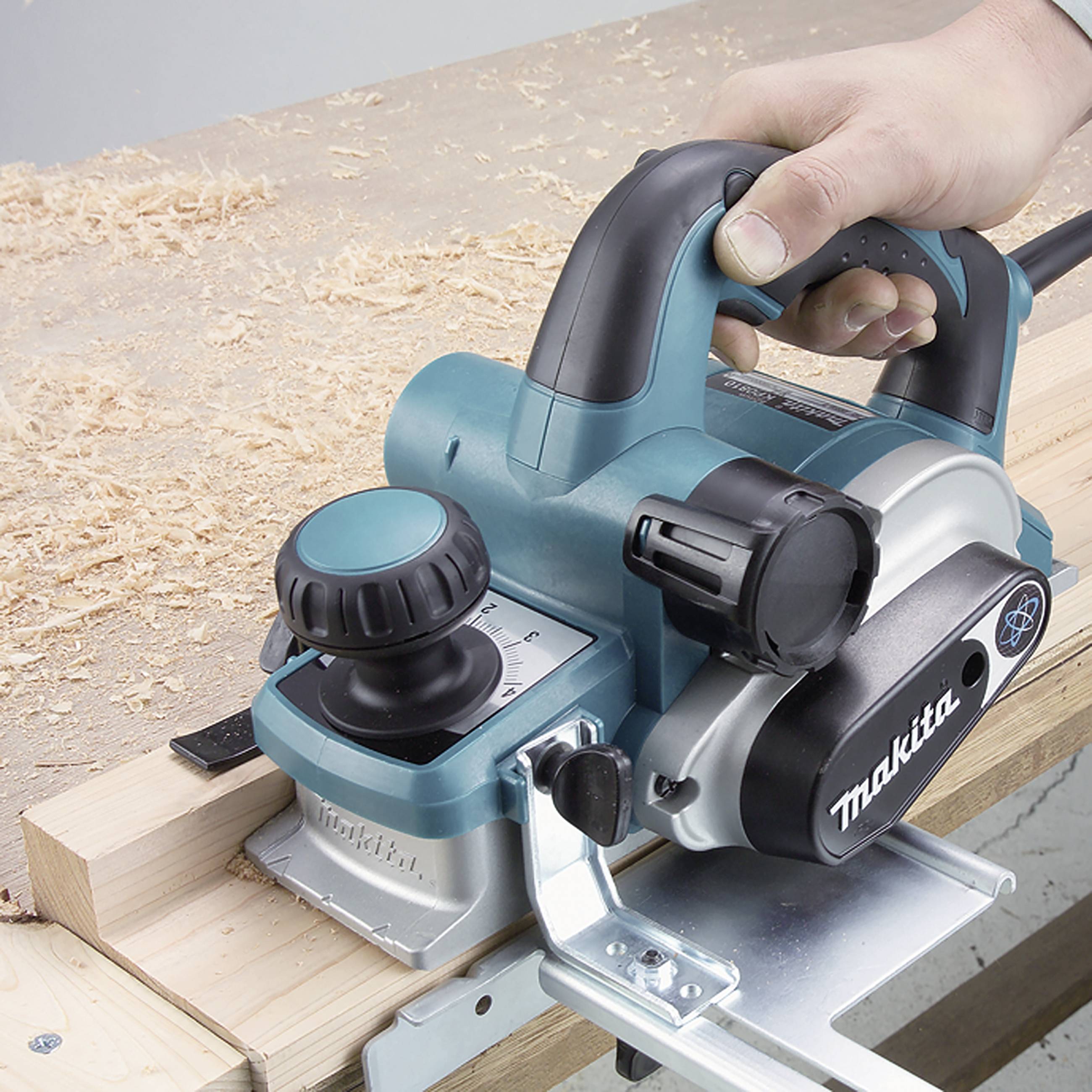 A hand guides a blue electric planer over a wooden surface, with wood shavings in the foreground.