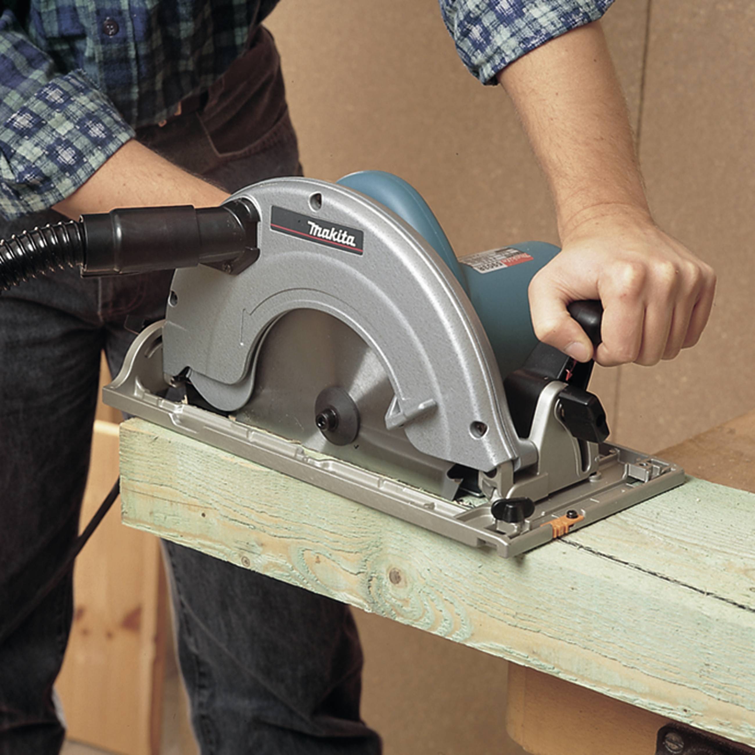 A person is using a handheld circular saw to cut a wooden board. The blue saw is connected to a dust extraction system.
