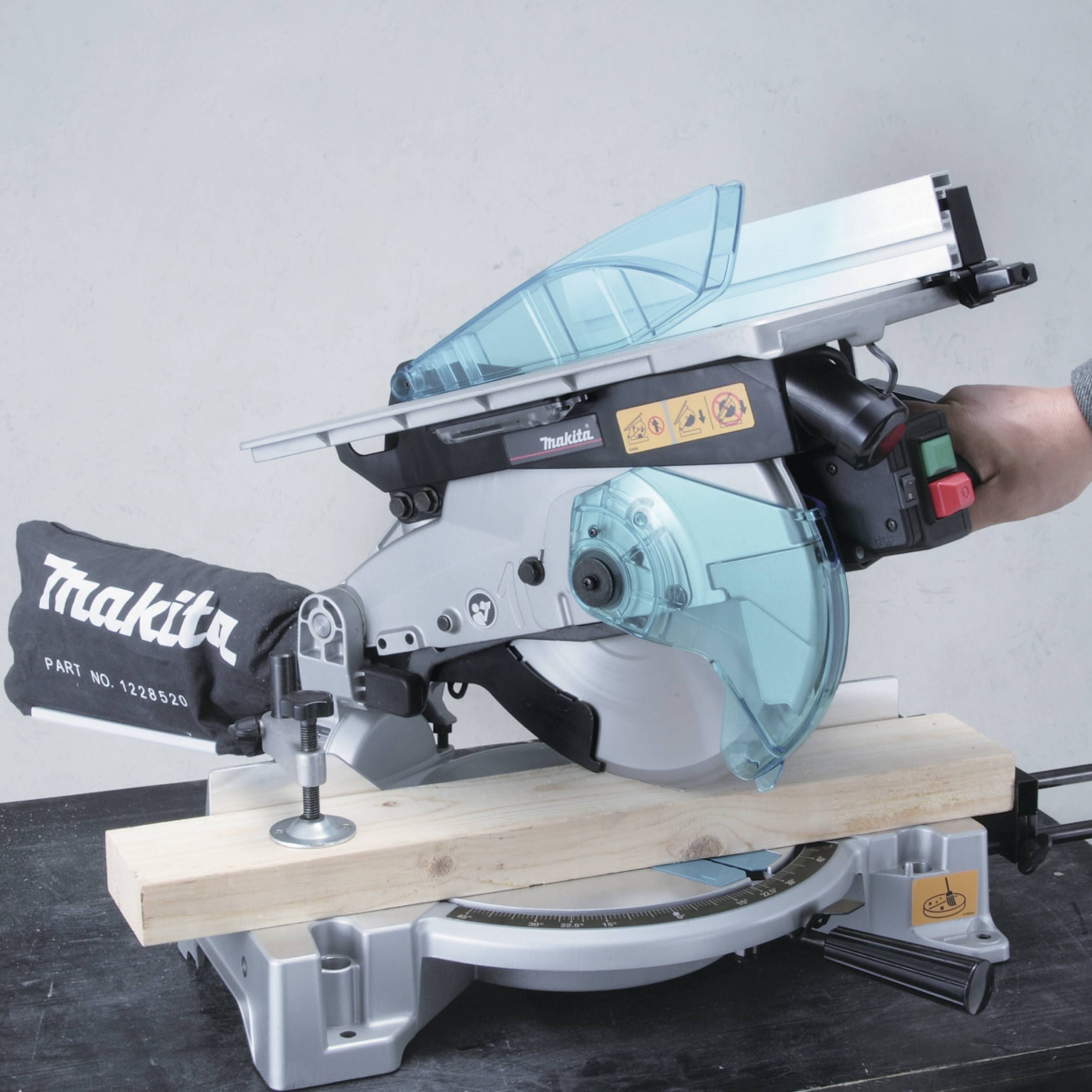 'A Makita mitre saw on a table, cutting a wooden beam. A hand is operating the green power button.'