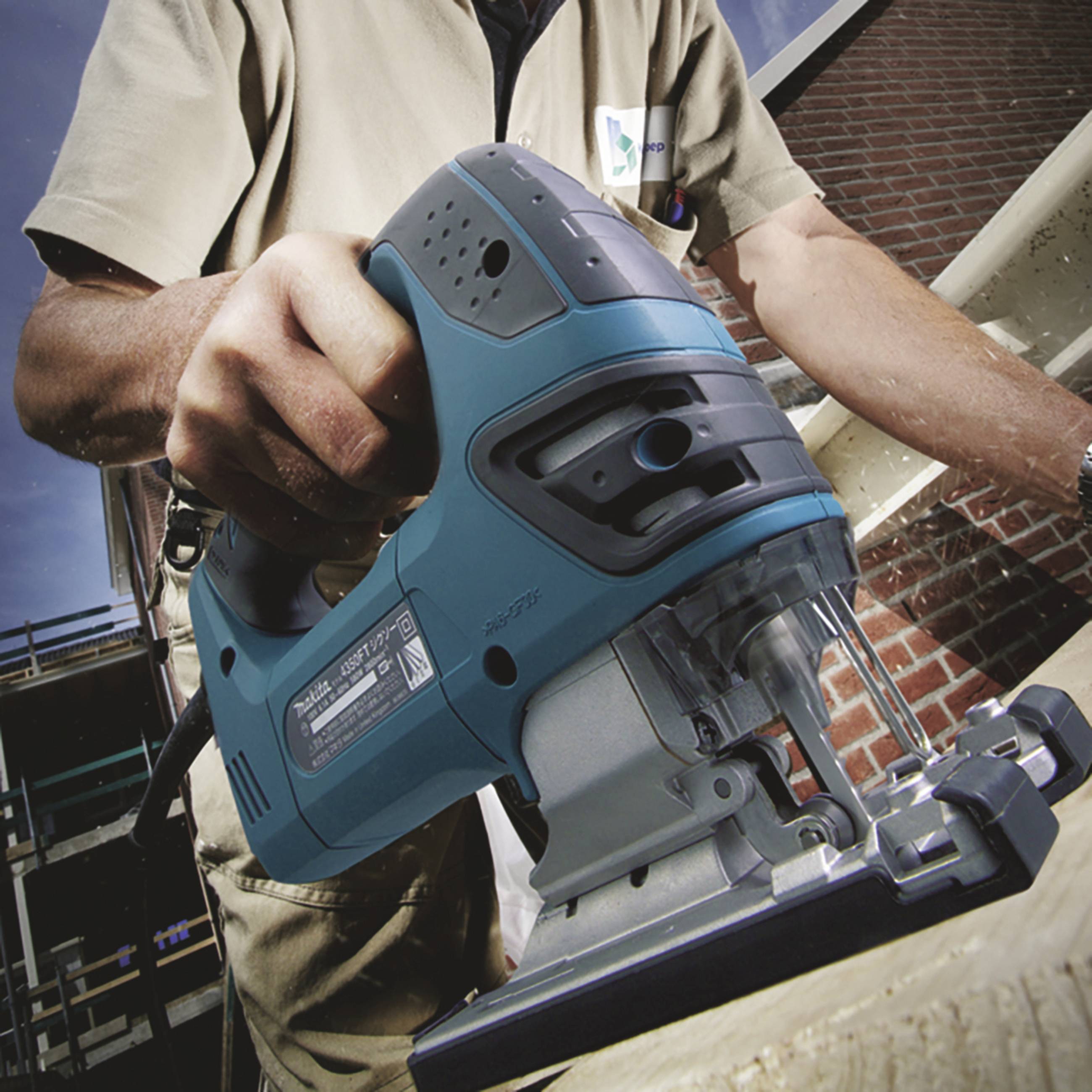 A person is holding an electric jigsaw and cutting through a wooden board. The person is wearing work clothes.