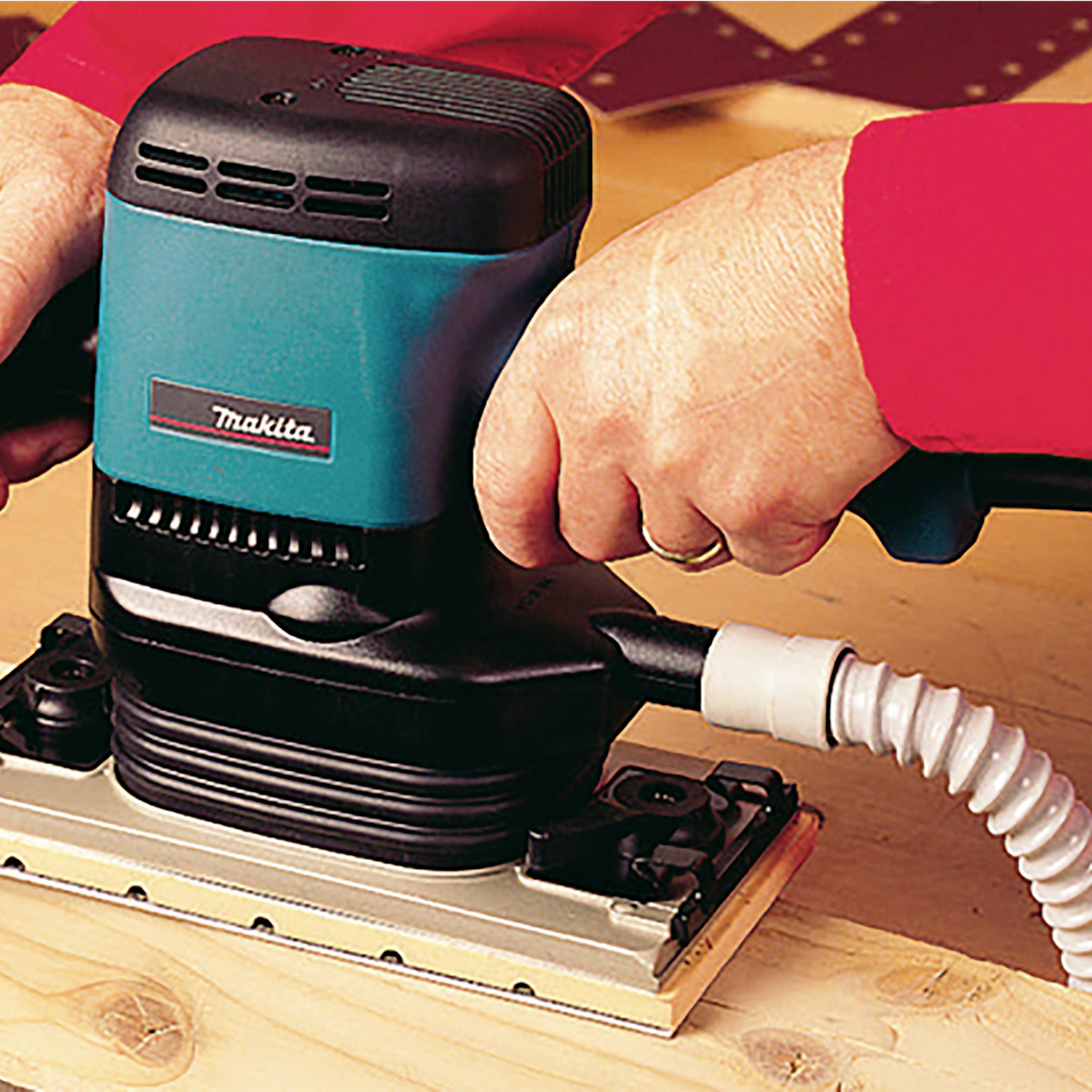 Two hands are holding a Makita electric orbital sander while working with wood.