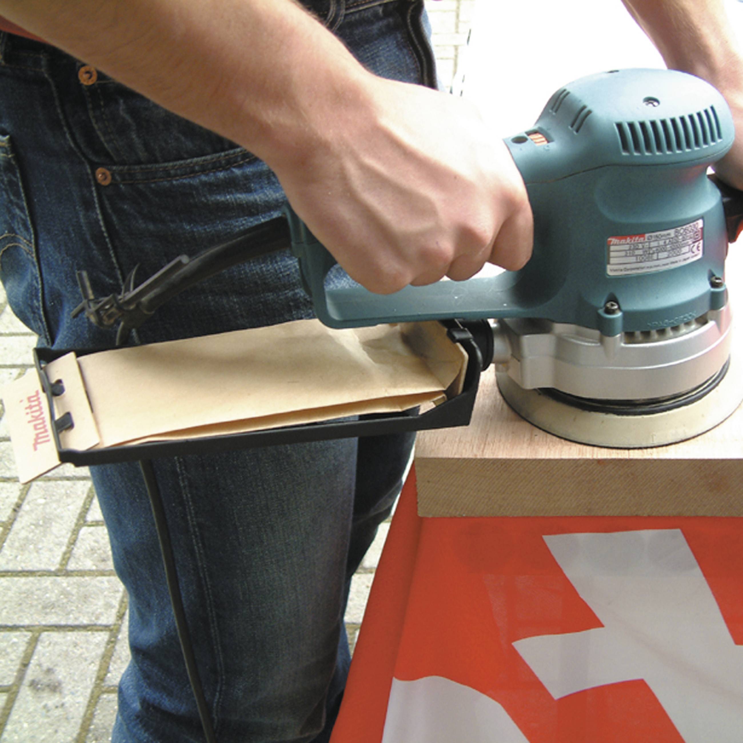 A person is sanding a piece of wood with a sanding machine. The focus is on the machine in action. Paving stones are visible in the background.