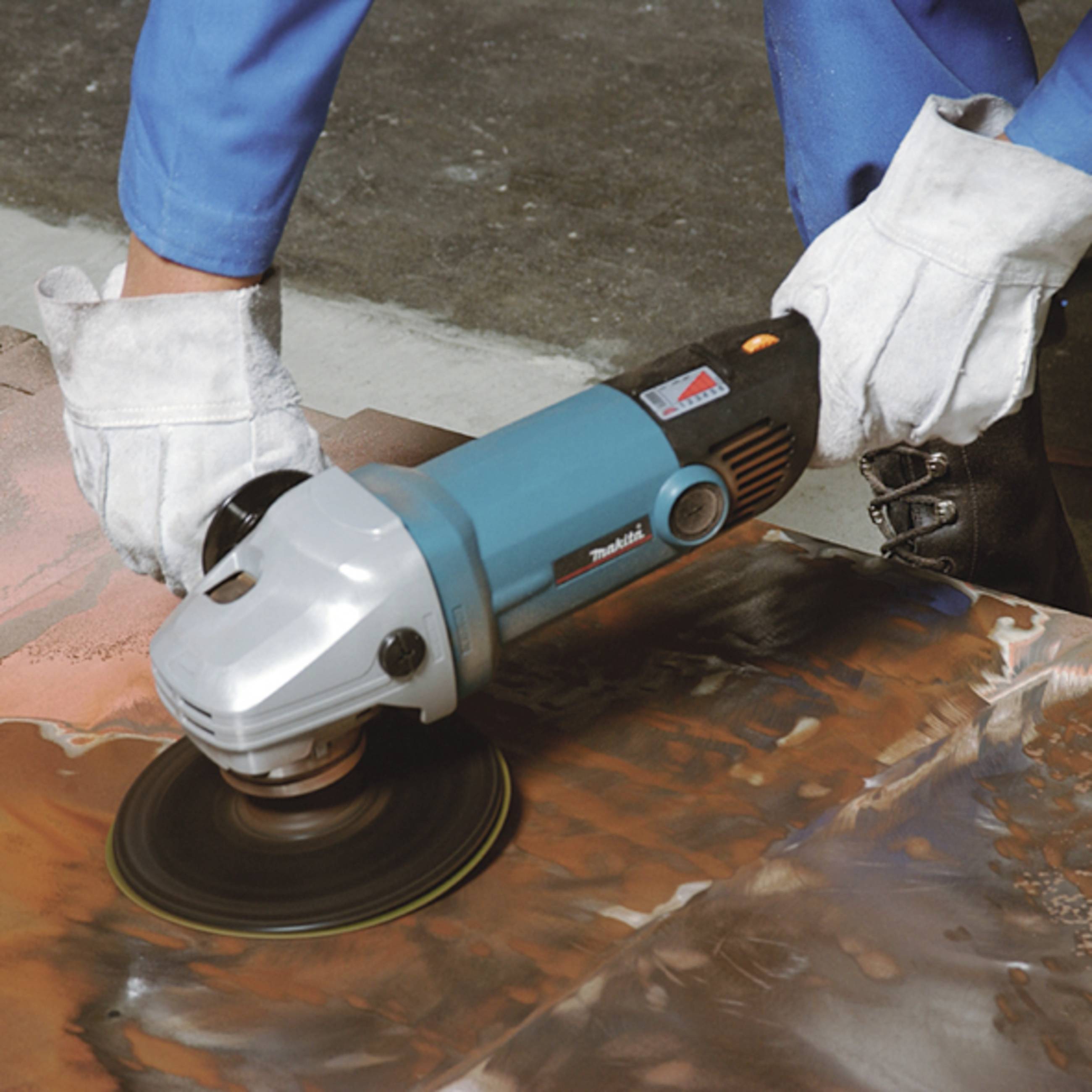 A person is grinding a metal surface with an angle grinder, wearing blue work trousers and white gloves, with a concrete floor in the background.