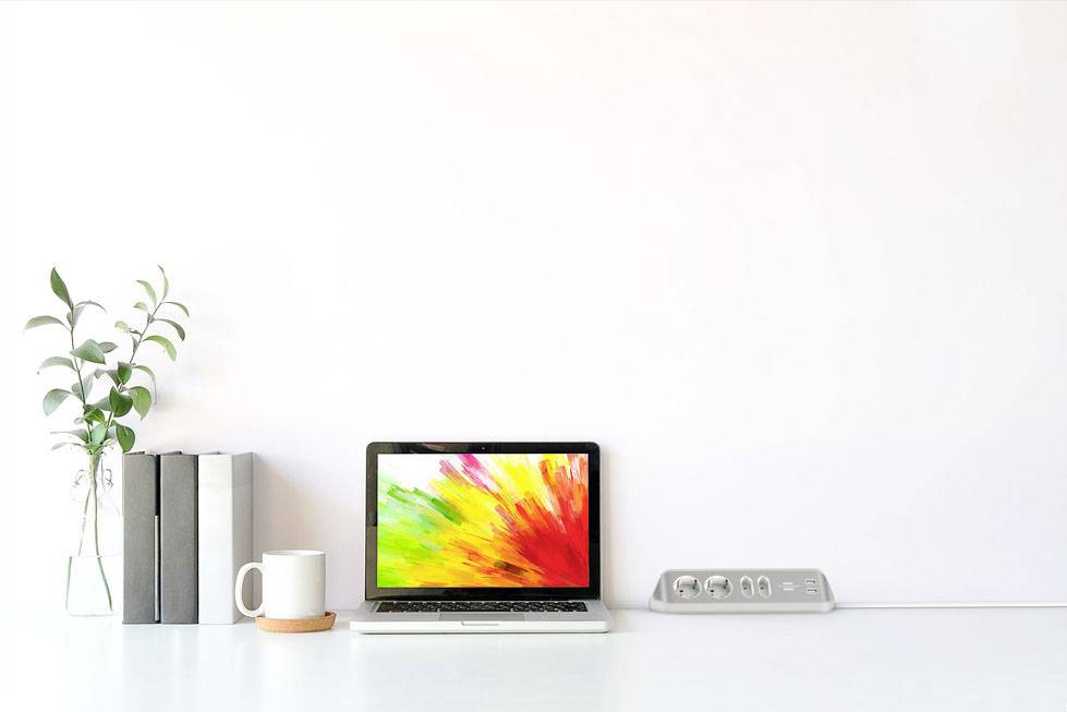 A laptop with a coloured screen sits on a white desk, surrounded by books, a plant, and a mug.