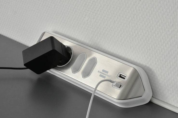 A multi-socket extension lead mounted on a wall with a black plug adapter inserted and a white charging cable connected.