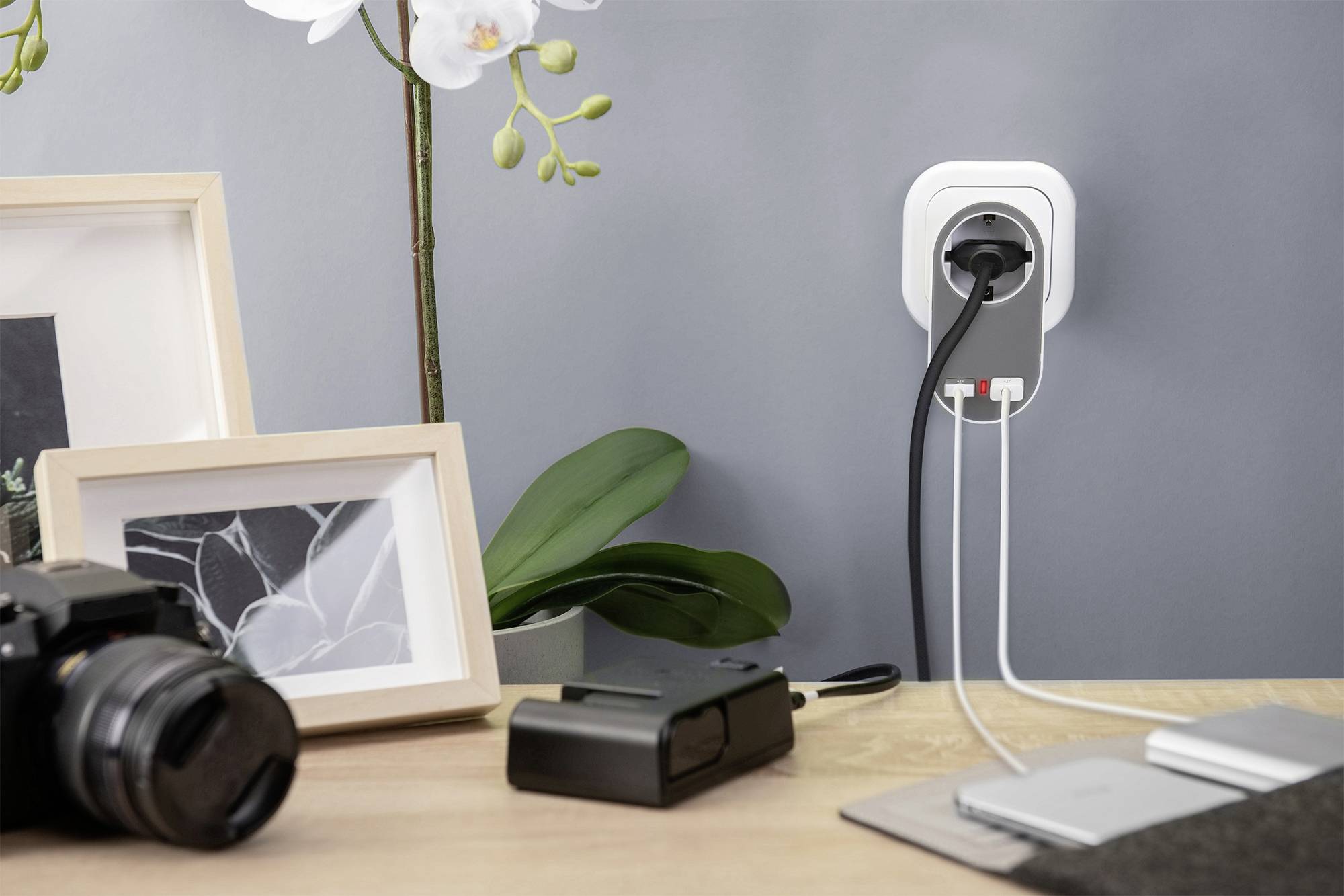 Three-pin socket with three connected cables on a wall. A camera and a picture frame are sitting on the table beside it.