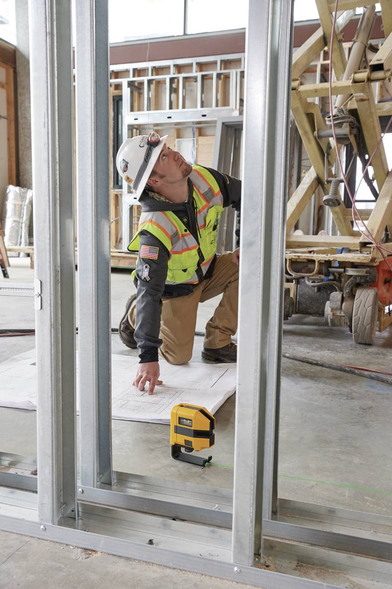 A construction worker wearing a hard hat and high-visibility vest is kneeling on a building site, checking construction plans next to a laser measuring device.
