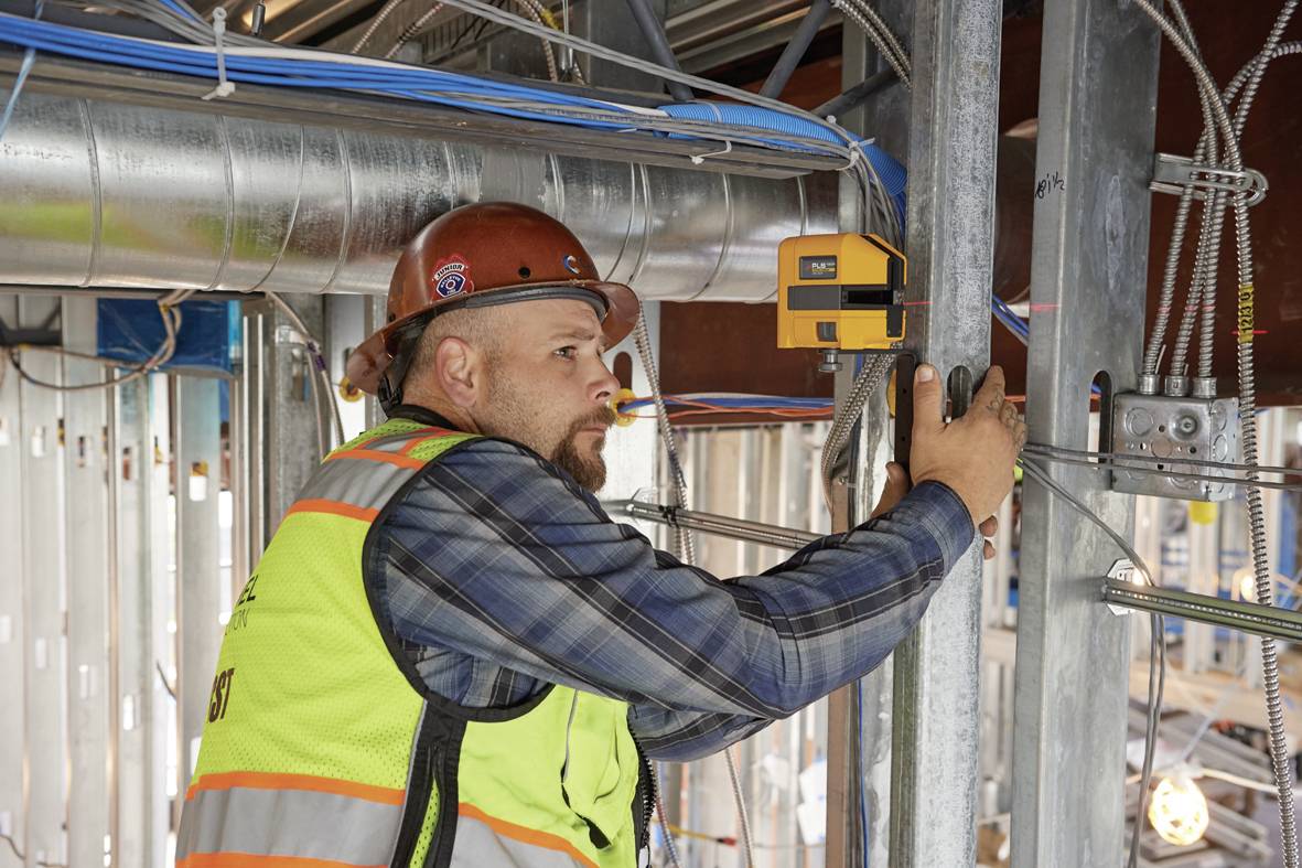 A construction worker in protective gear is using a measuring device in an unfinished building. Electrical cables and pipes are visible.